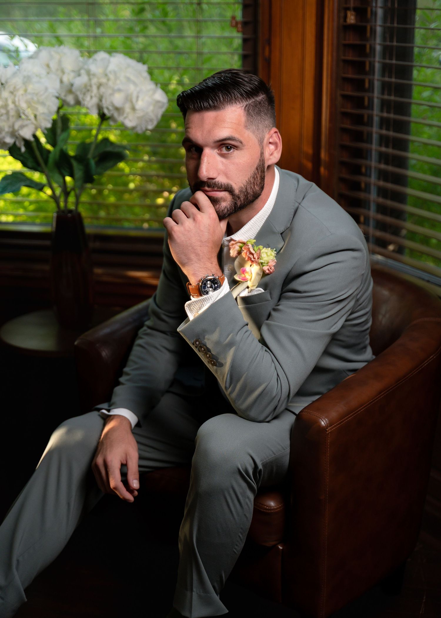 Man in gray suit with floral boutonniere sits thoughtfully in leather chair near white hydrangeas and wooden blinds.