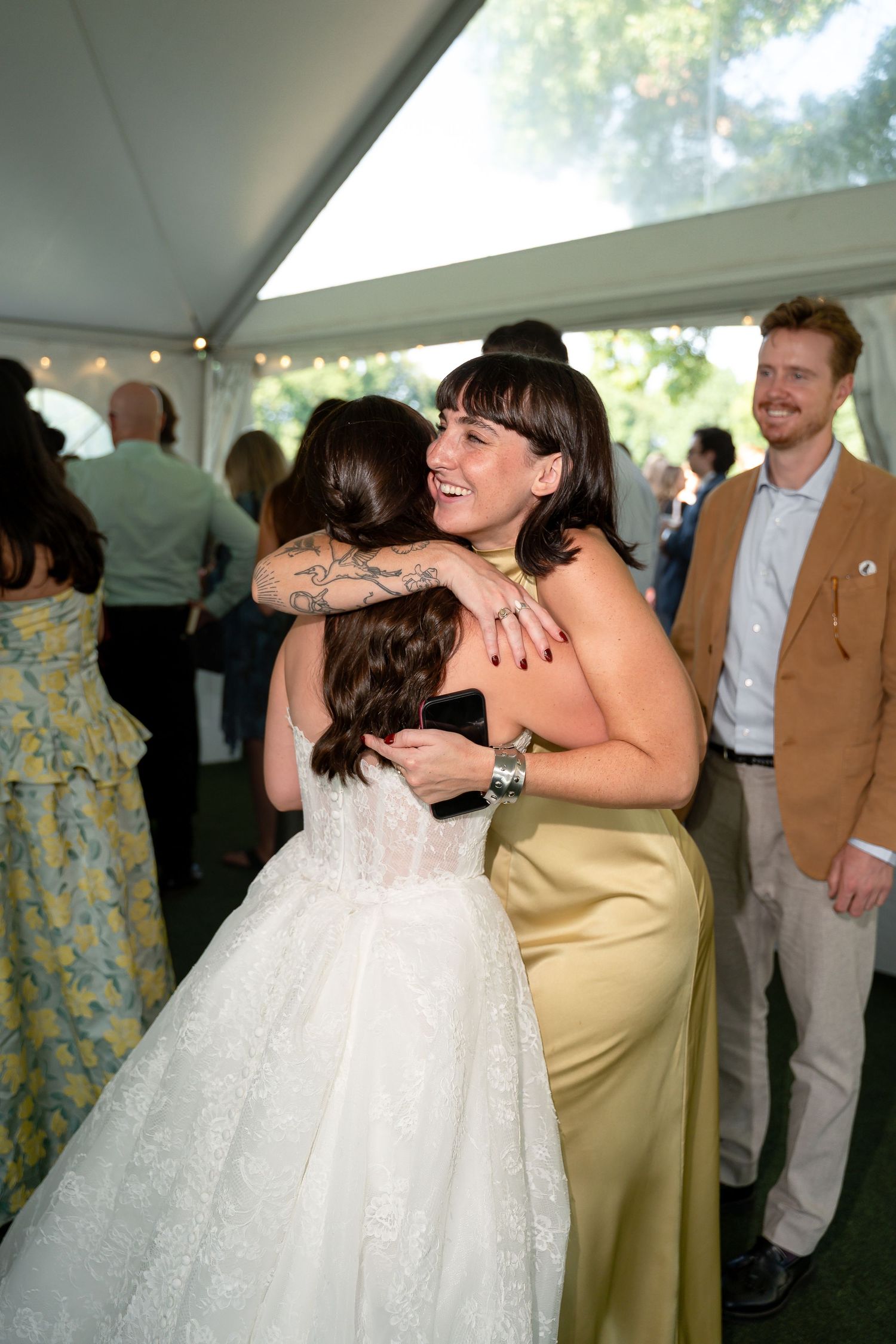 Bride in white lace gown hugging a guest in gold satin dress at an outdoor wedding reception under a tent.