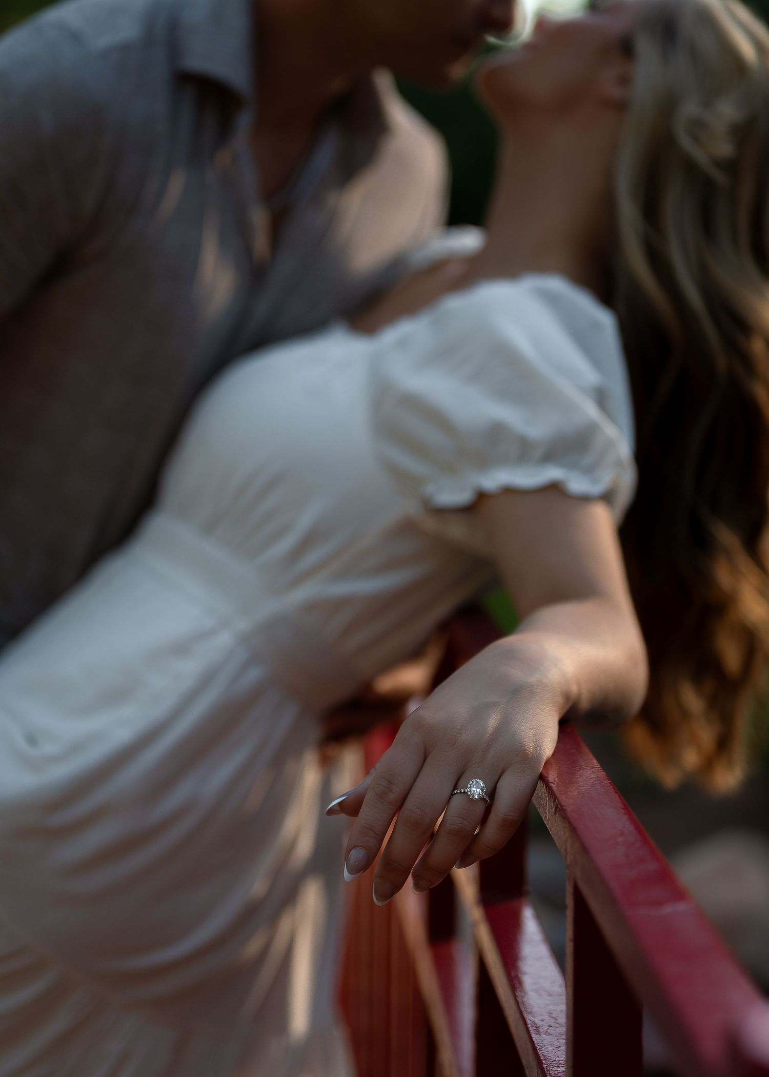 Couple sharing a romantic kiss on a bridge, woman in white dress displaying a diamond engagement ring on the railing.