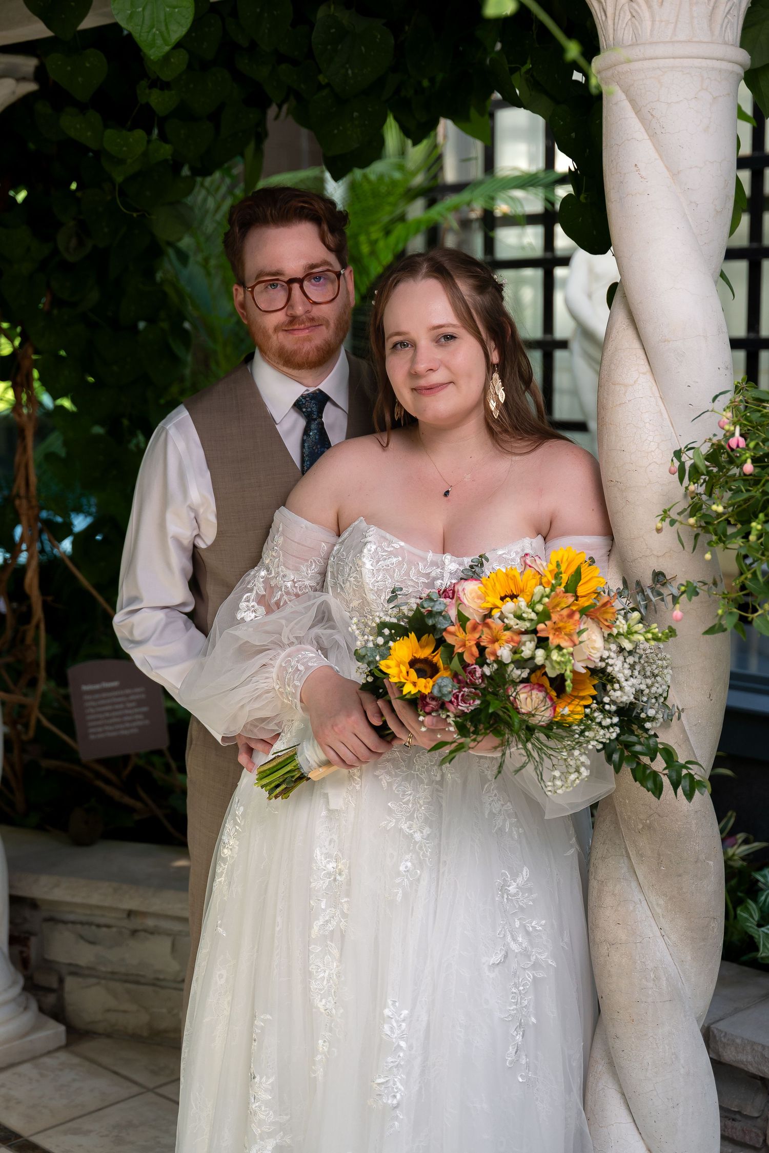 Bride in white lace gown holding sunflower bouquet poses with groom in vest and tie near white columns.