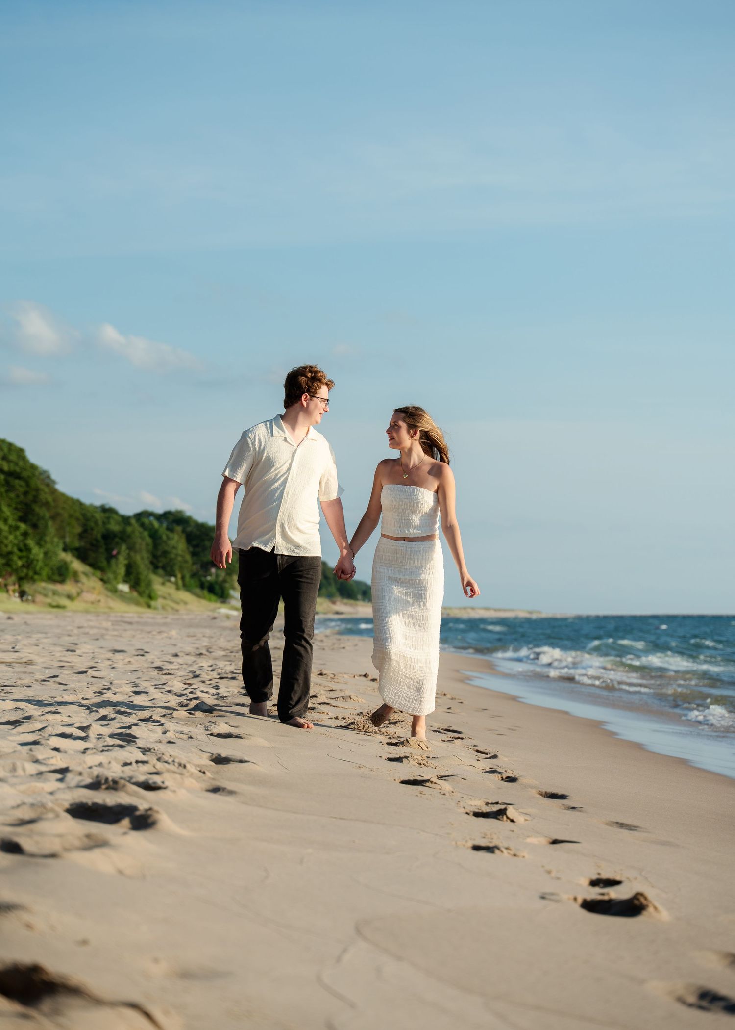 Couple walking hand in hand along a beach leaving footprints in the sand with ocean waves nearby.