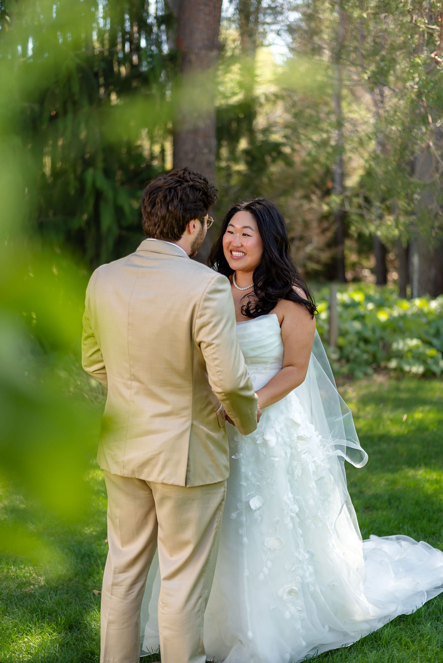 Smiling bride in white floral gown holds hands with groom in tan suit outdoors in lush green garden setting.