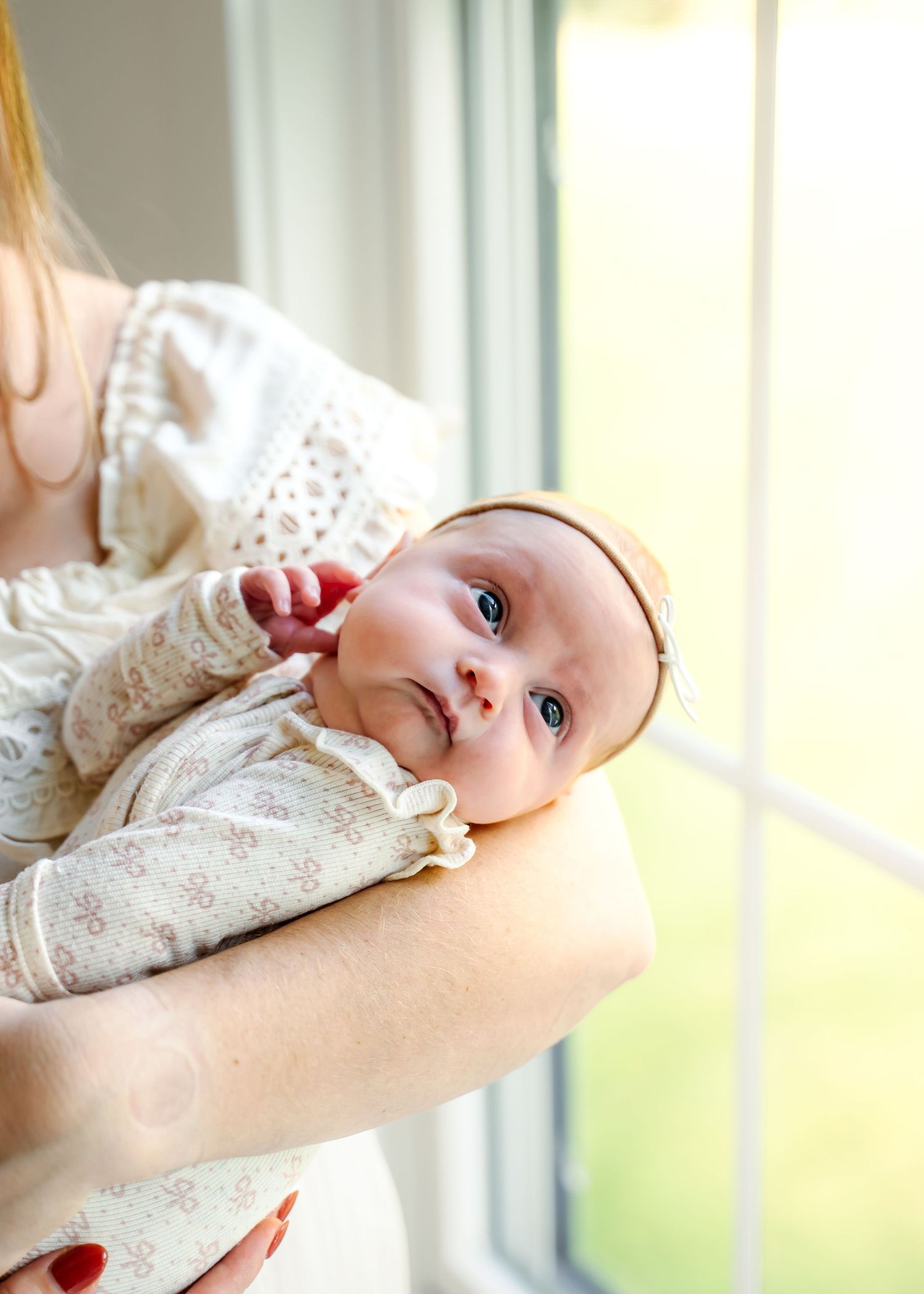 A tiny baby in a white lace dress lies gently cradled in someone's arms near a window.