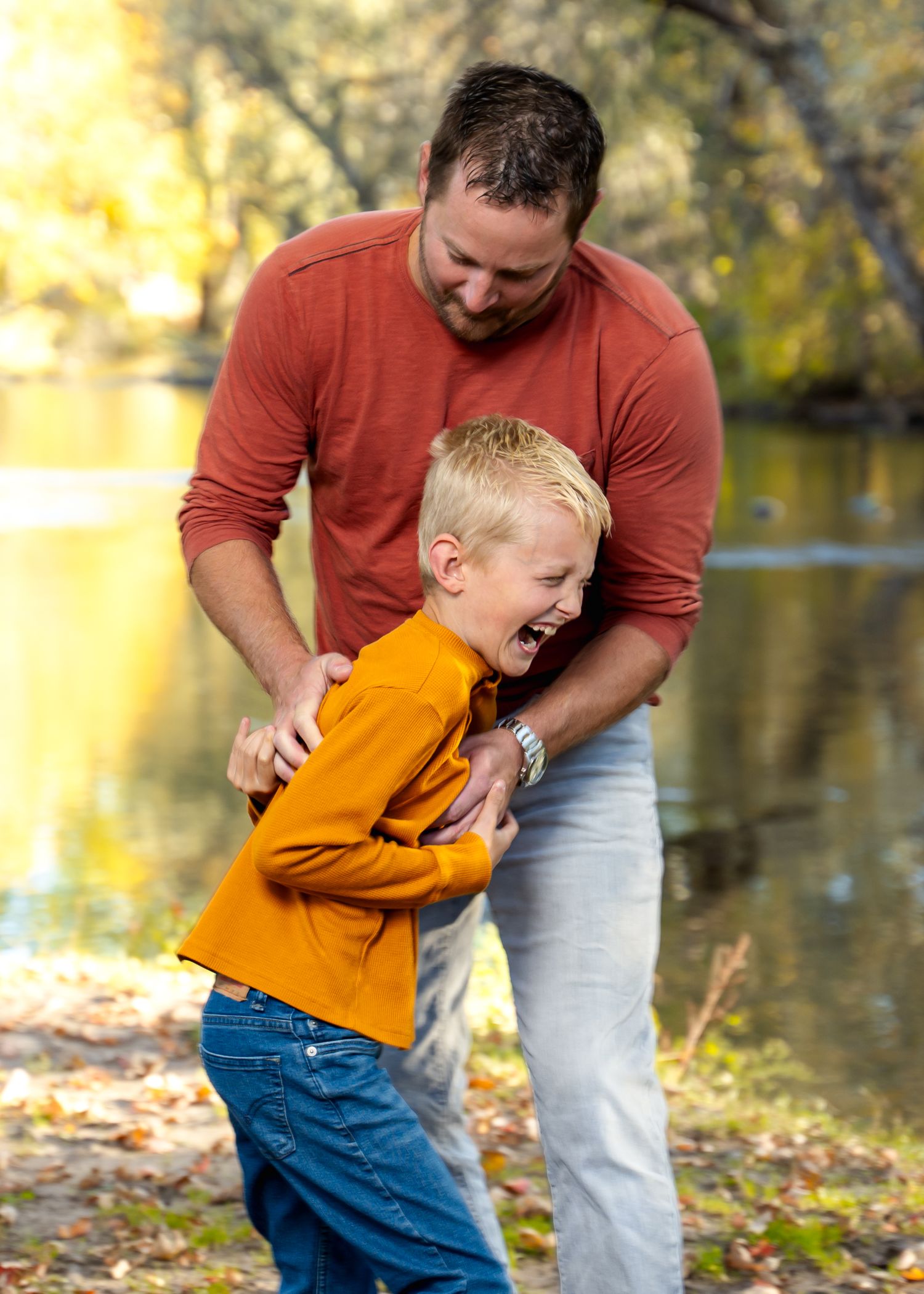 Playful tickling moment between parent and child in orange shirt by riverside.