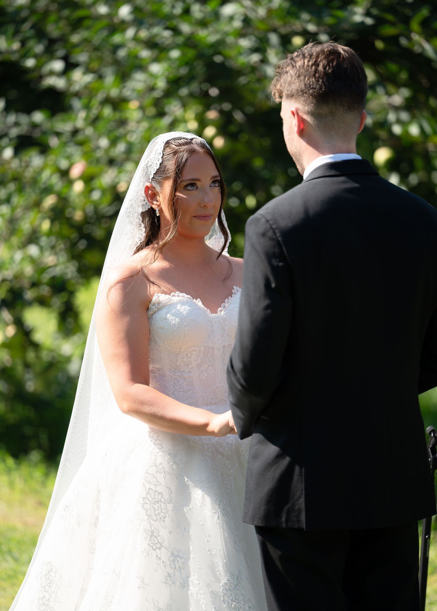 Bride in white lace gown and veil exchanges vows with groom in black suit during outdoor wedding ceremony.