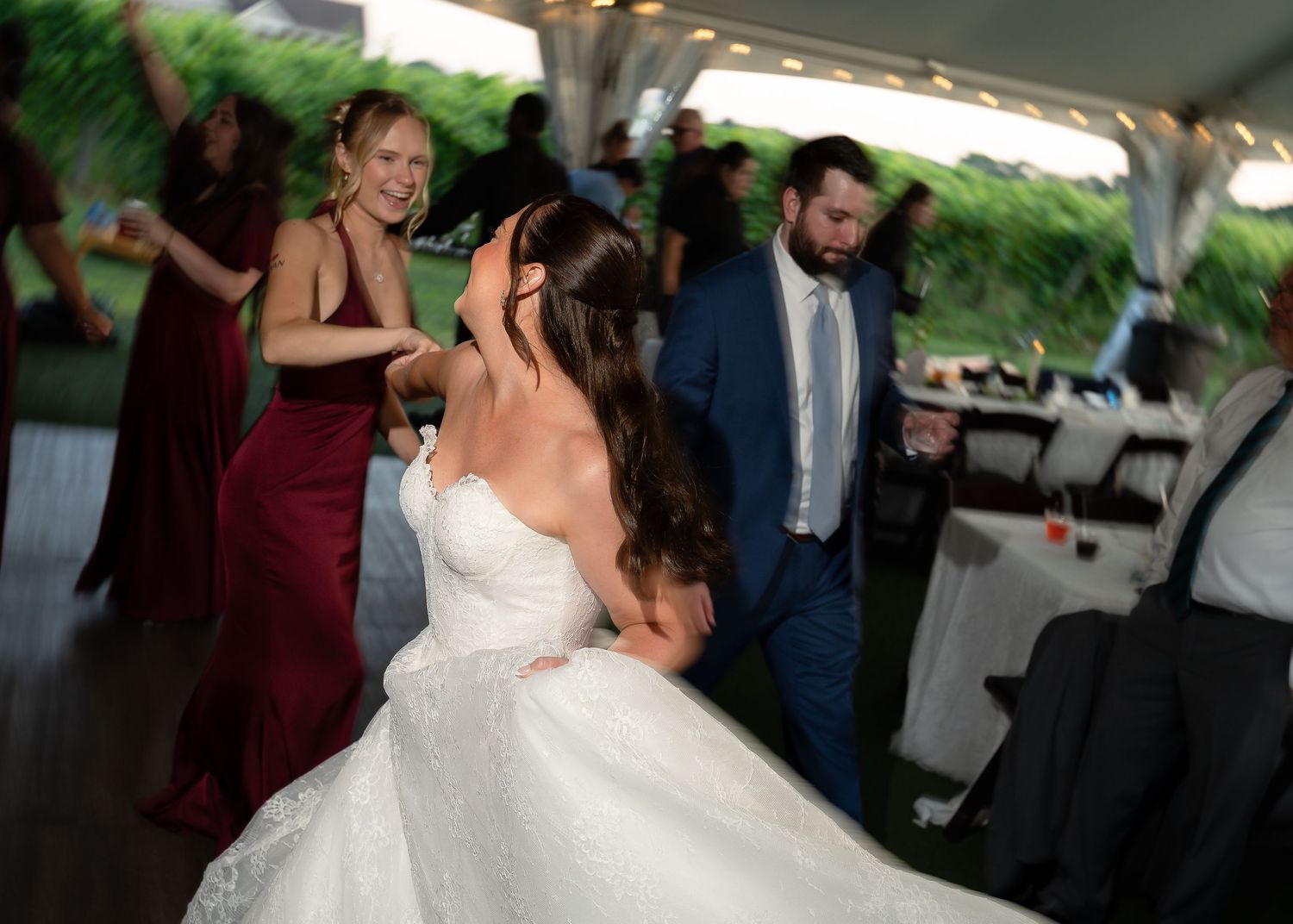 Bride in white gown dancing with guests at outdoor wedding reception under tent with bridesmaids in burgundy dresses.