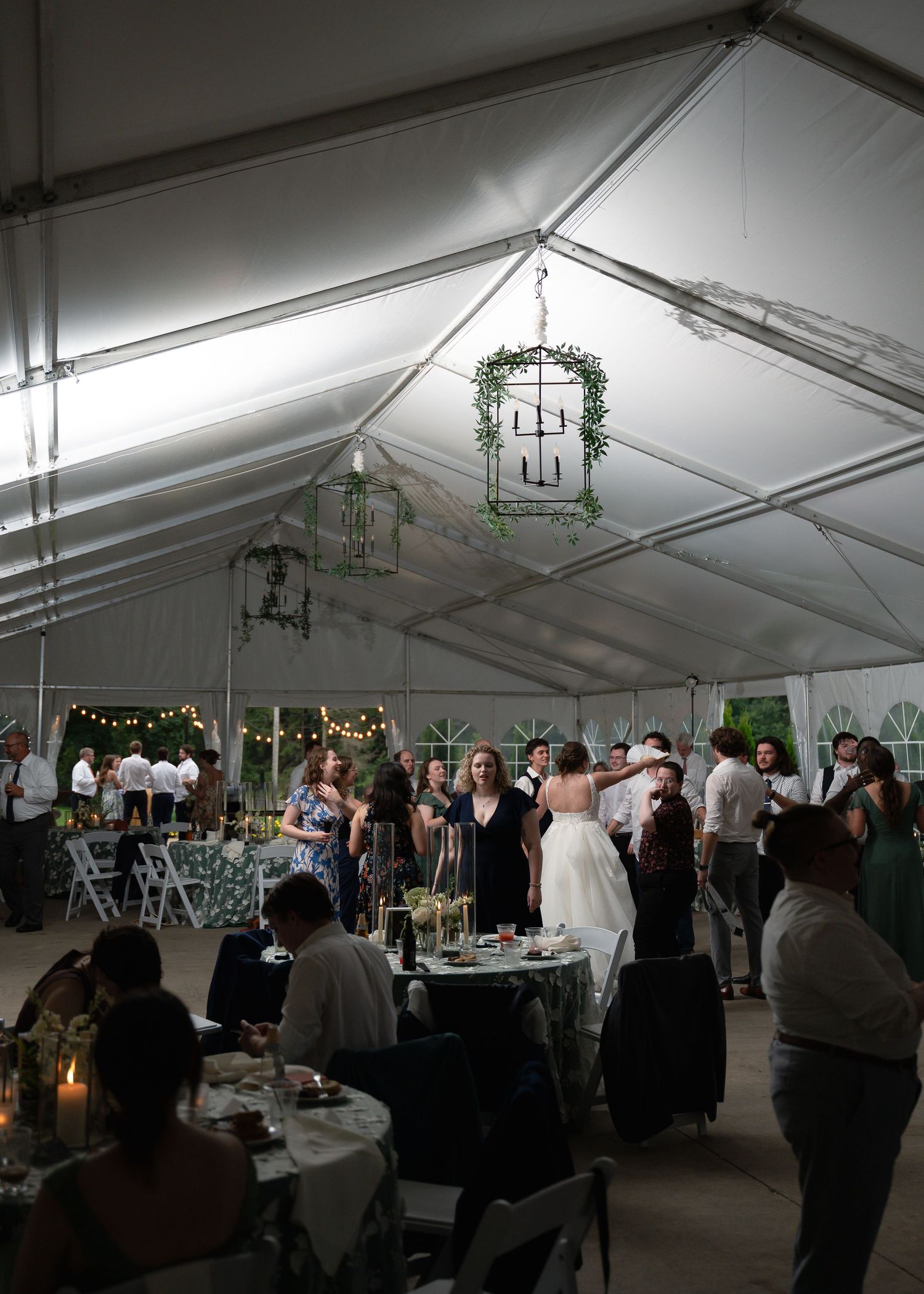 Guests mingle and dance under a white tent adorned with greenery chandeliers at an elegant outdoor wedding reception.