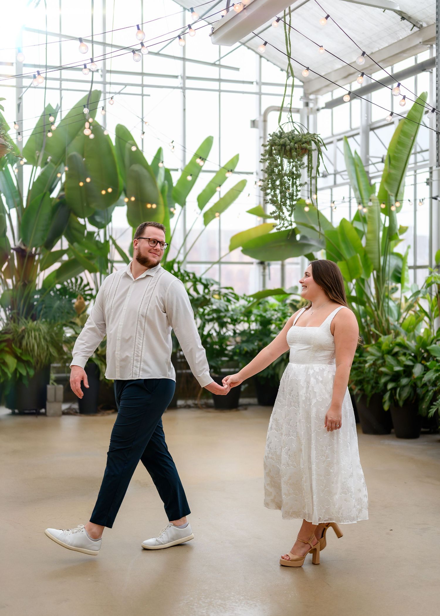 Couple dancing and laughing together in a bright greenhouse filled with tropical plants and hanging baskets.