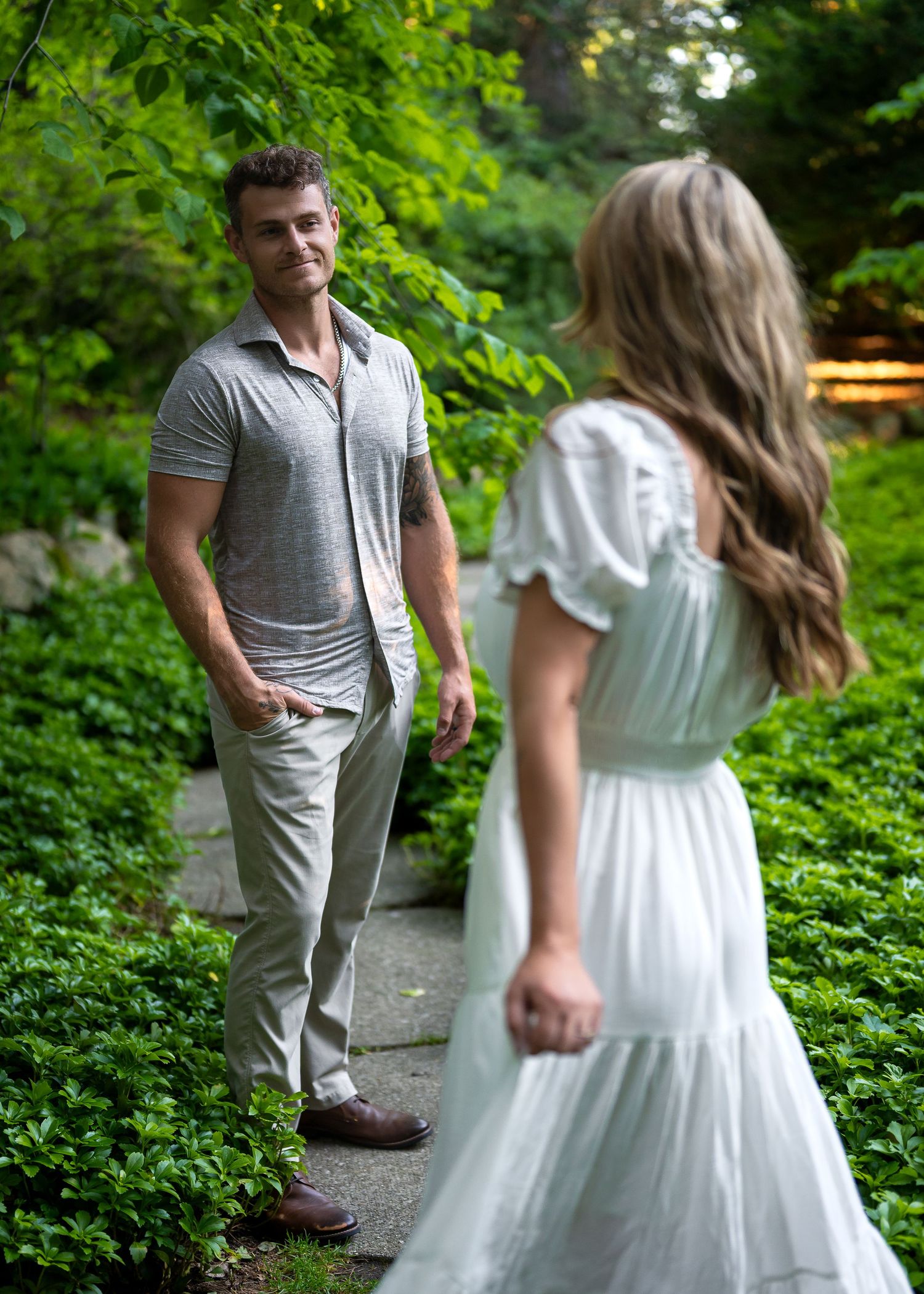 A man in a gray polo and khakis stands on a garden path, smiling at a woman in a white dress.