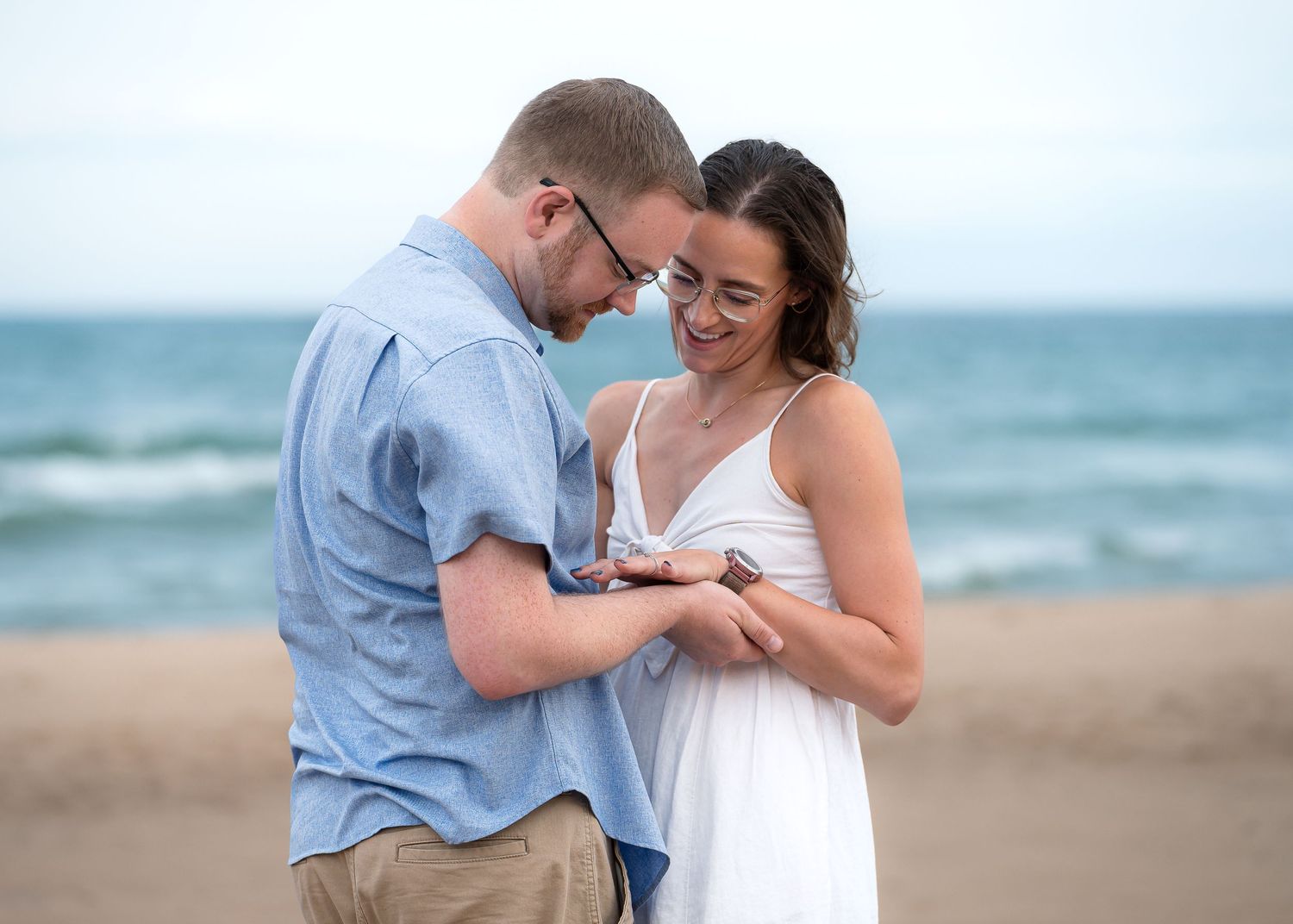 Couple sharing a tender moment on the beach, examining an engagement ring, with ocean waves in the background.