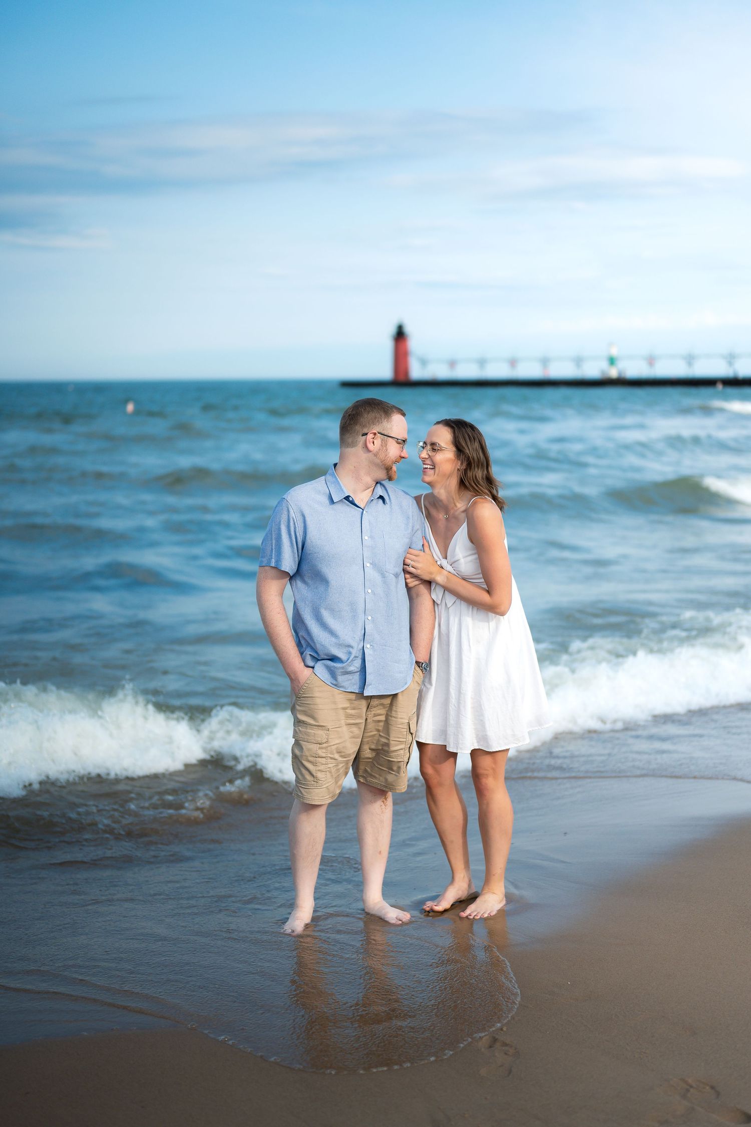 Couple standing barefoot on sandy beach shore with ocean waves and red lighthouse in background.