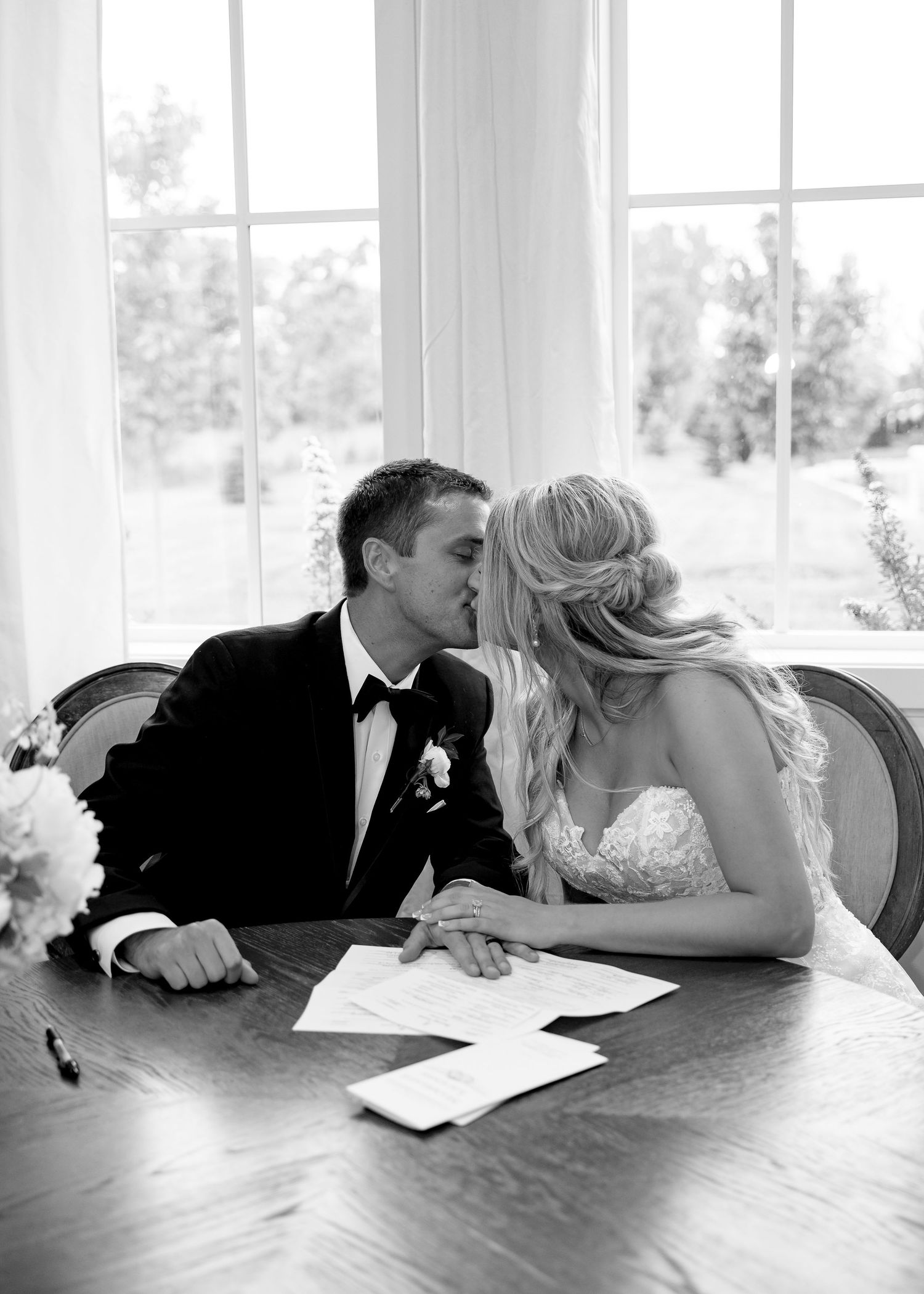 Black and white photo of a bride and groom sharing a kiss while signing their wedding marriage certificate at a table.