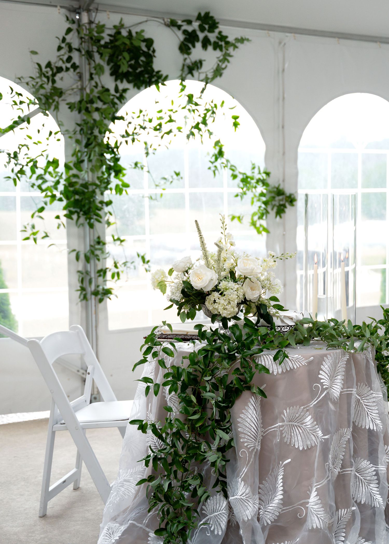 White tent wedding reception sweetheart table adorned with lush greenery garland and white floral centerpiece.