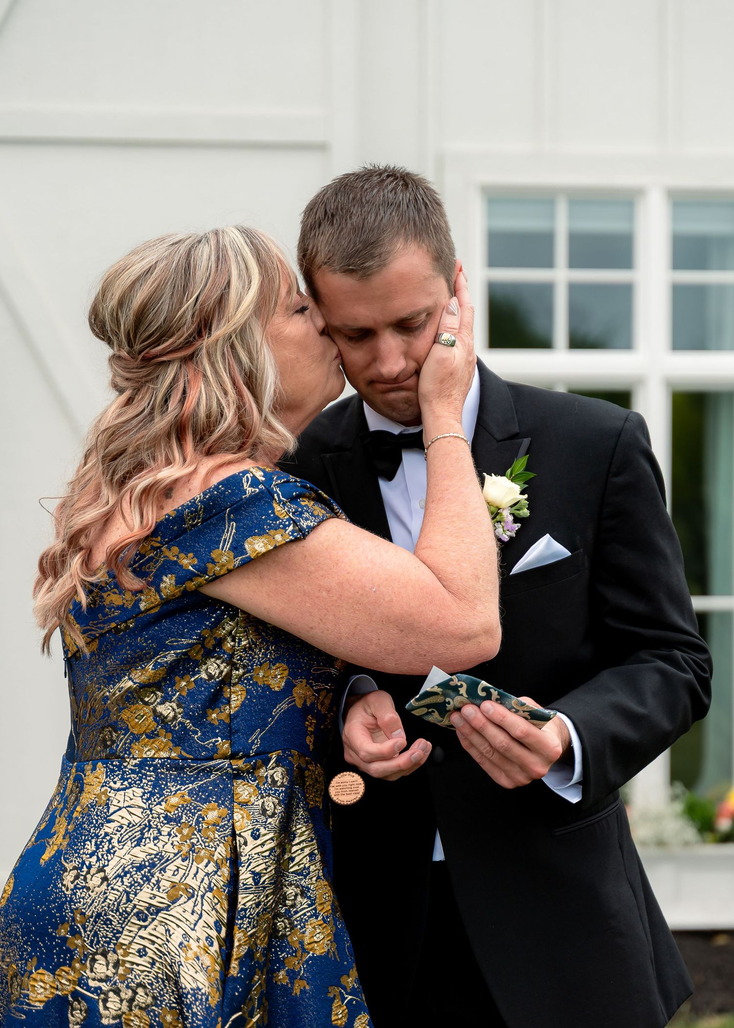 A woman in a blue and gold dress kisses a groom in a tuxedo on the cheek at a wedding.