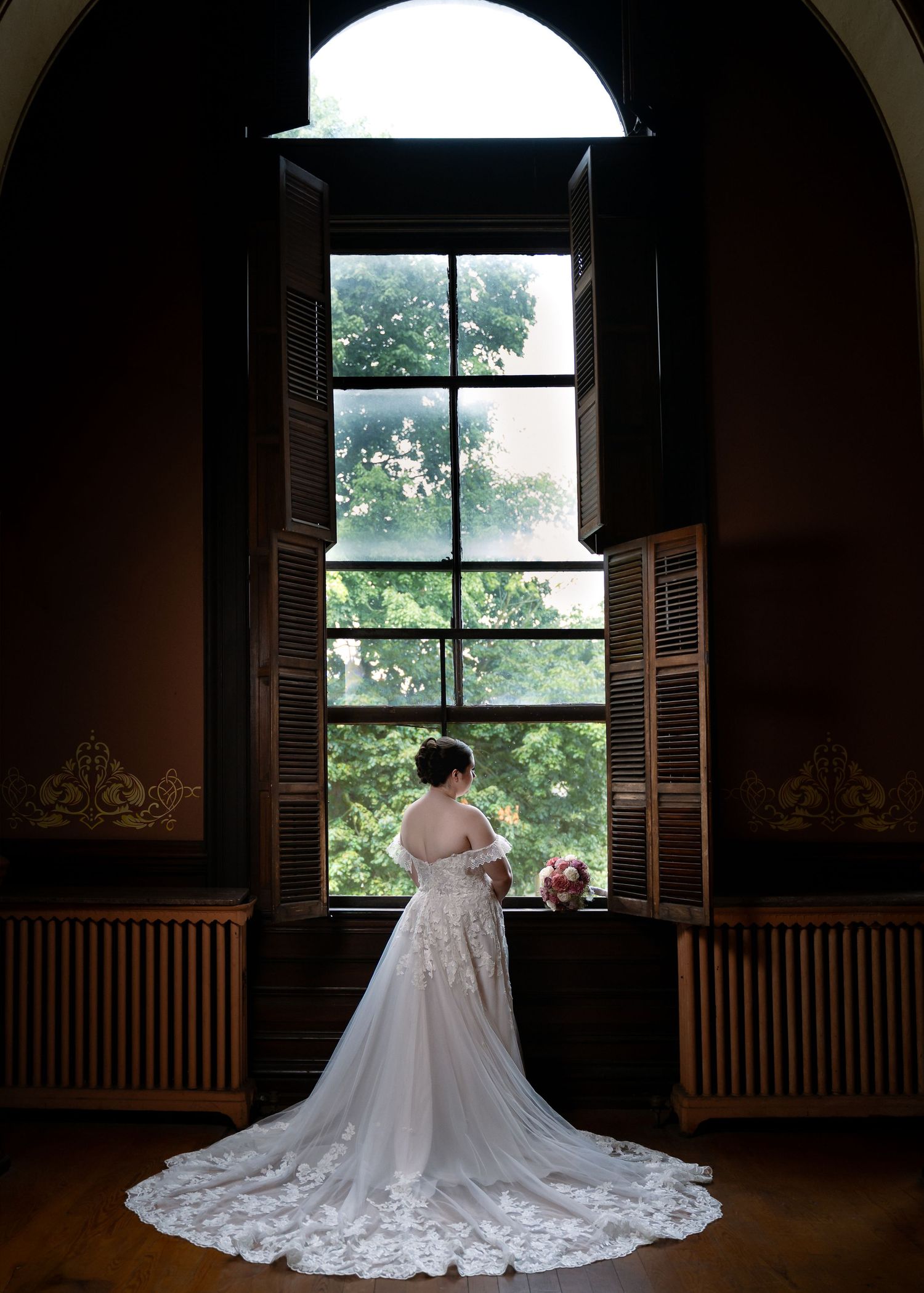 Bride in white lace wedding gown with long train stands before tall arched window in elegant historic venue.