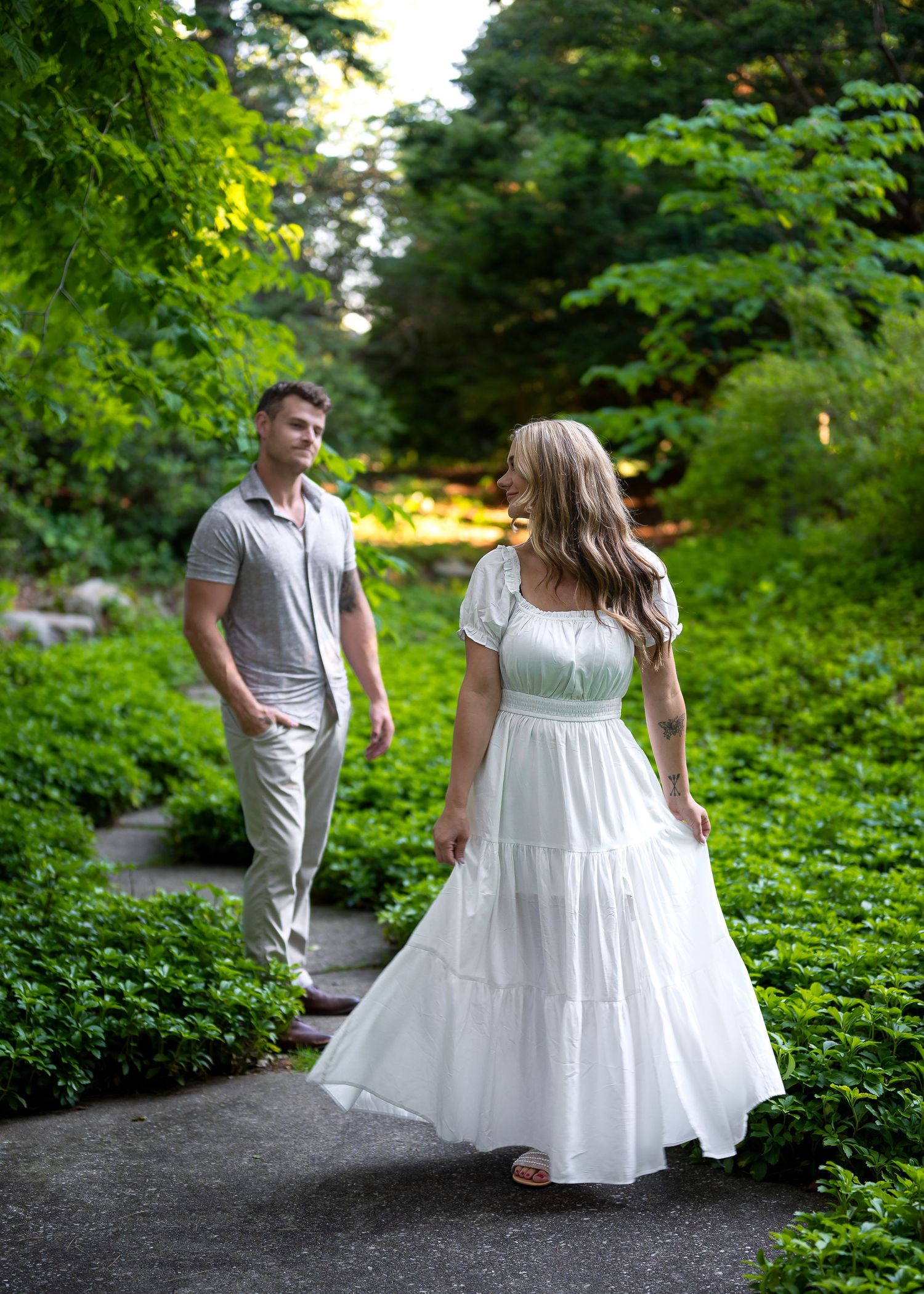 Couple walking on a garden path surrounded by lush green foliage, woman in white maxi dress looks back at man in gray.