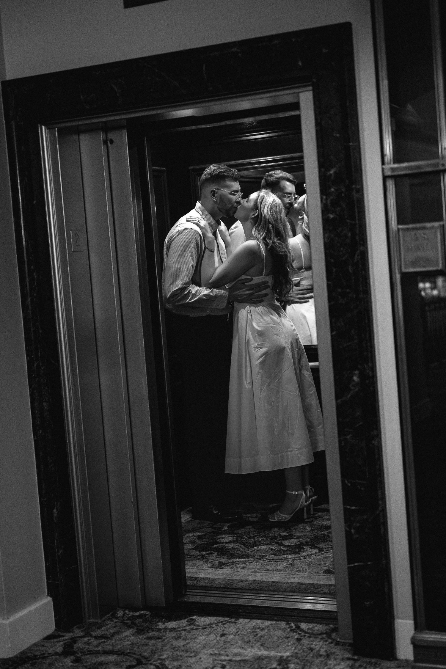 Black and white photo of a couple sharing a romantic kiss in an elegant elevator, reflected in mirrored walls.