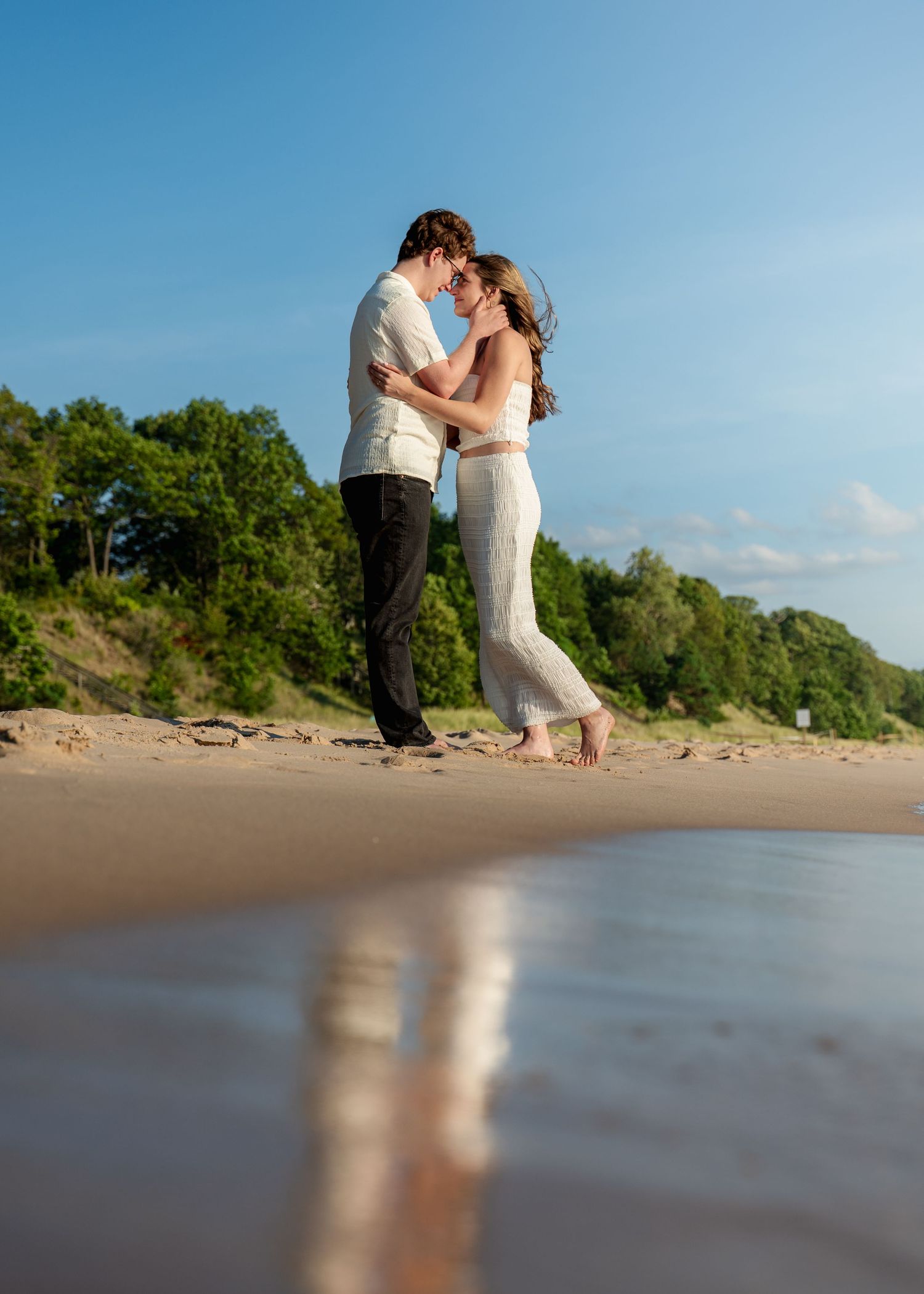 Romantic couple dancing together on a sandy beach with trees in the background during a sunny day.