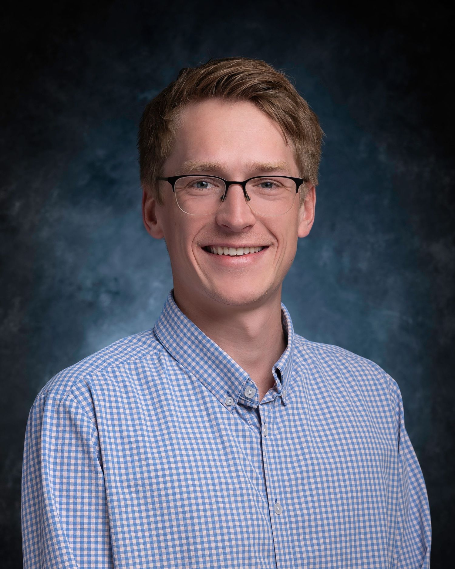Young man with glasses and light brown hair smiling, wearing a blue checkered shirt against a studio background.