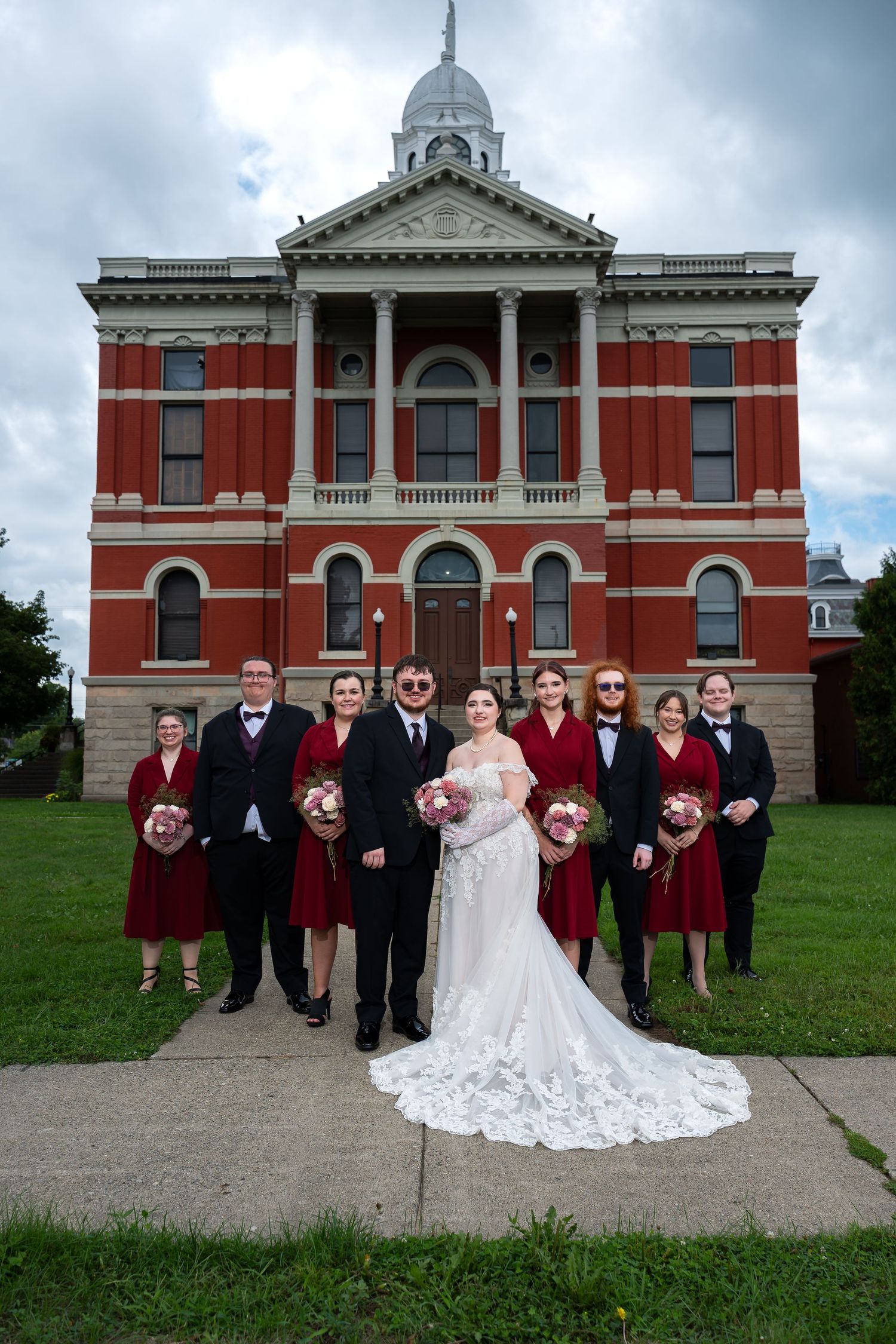 Wedding party poses before red brick historic courthouse, bride in white gown with bridesmaids in burgundy dresses.