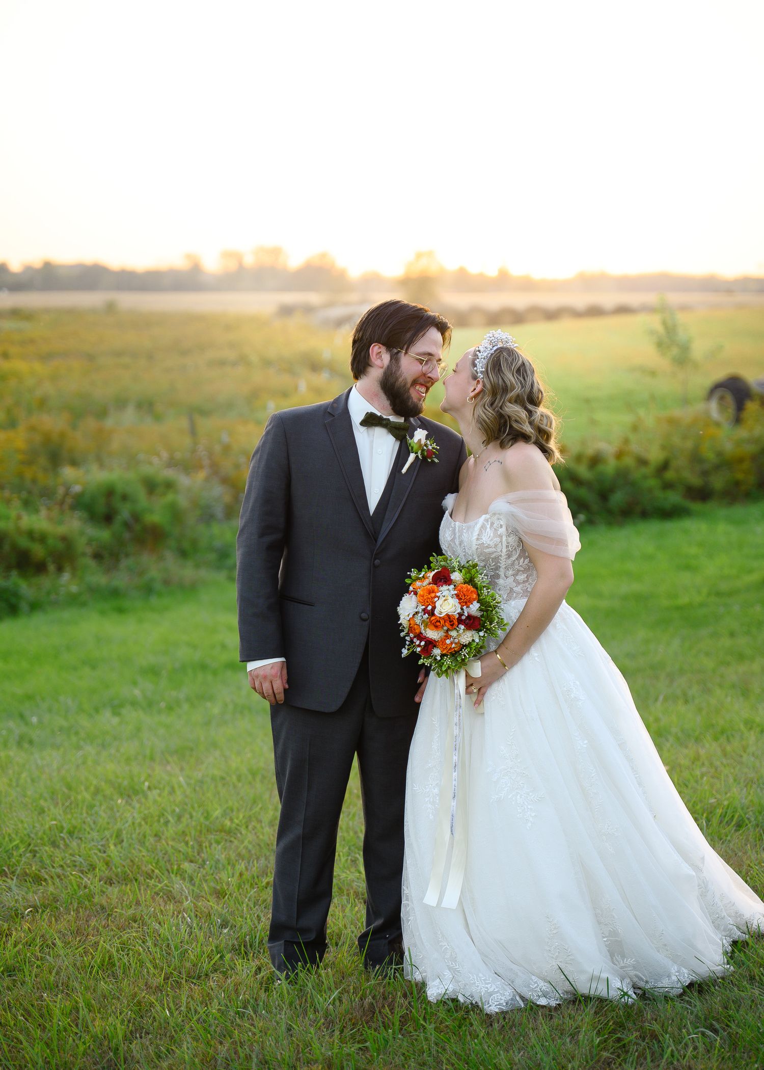 A wedding couple stands together in a golden field at sunset with the bride holding a colorful bouquet.