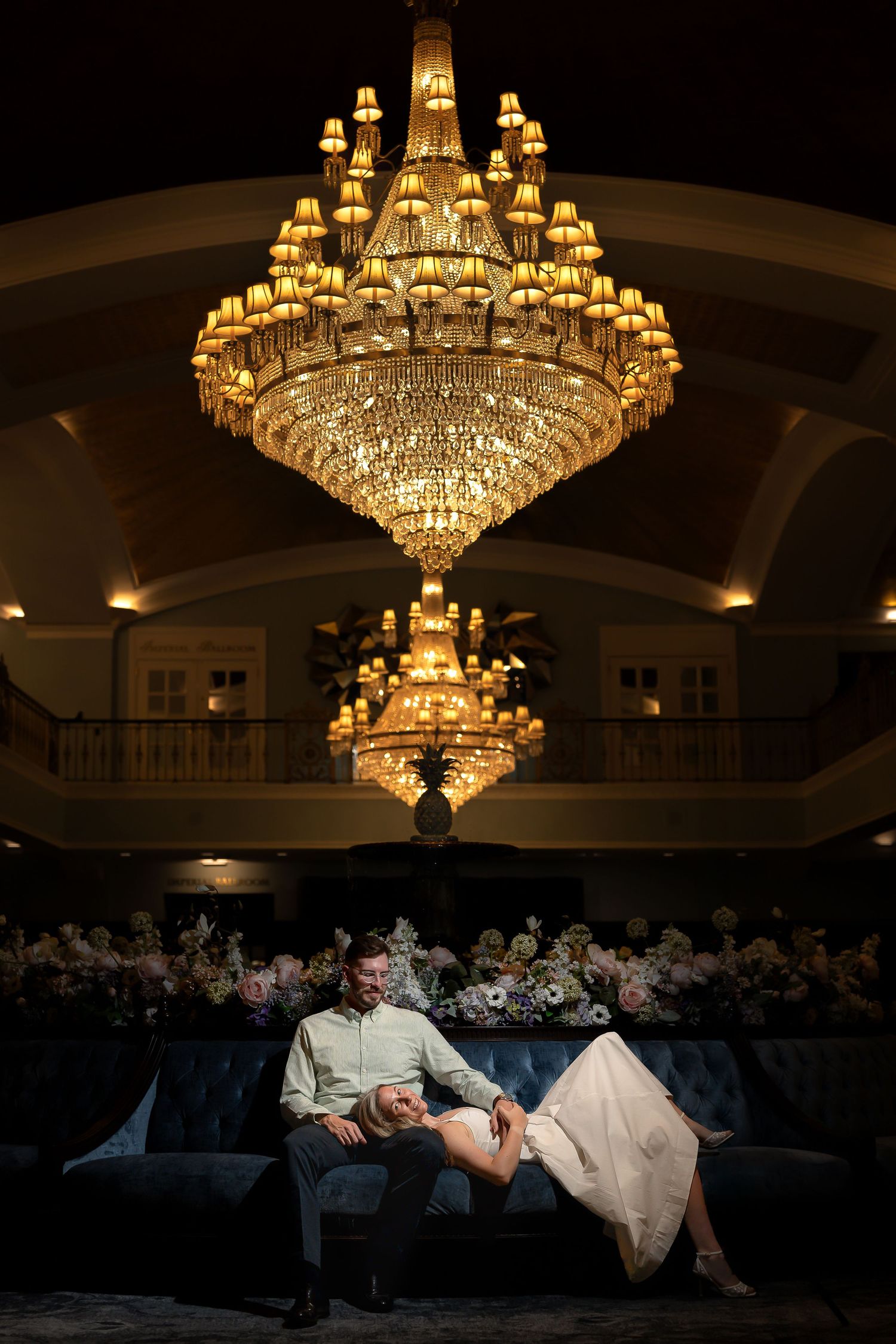 Couple sitting on velvet sofa beneath grand crystal chandelier in elegant ballroom with floral arrangements.