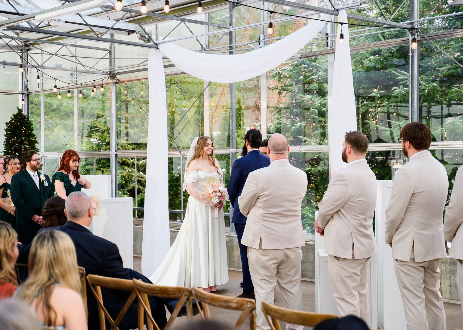Elegant greenhouse wedding ceremony with bride in white gown, groomsmen in tan suits, and draped white arch decor.