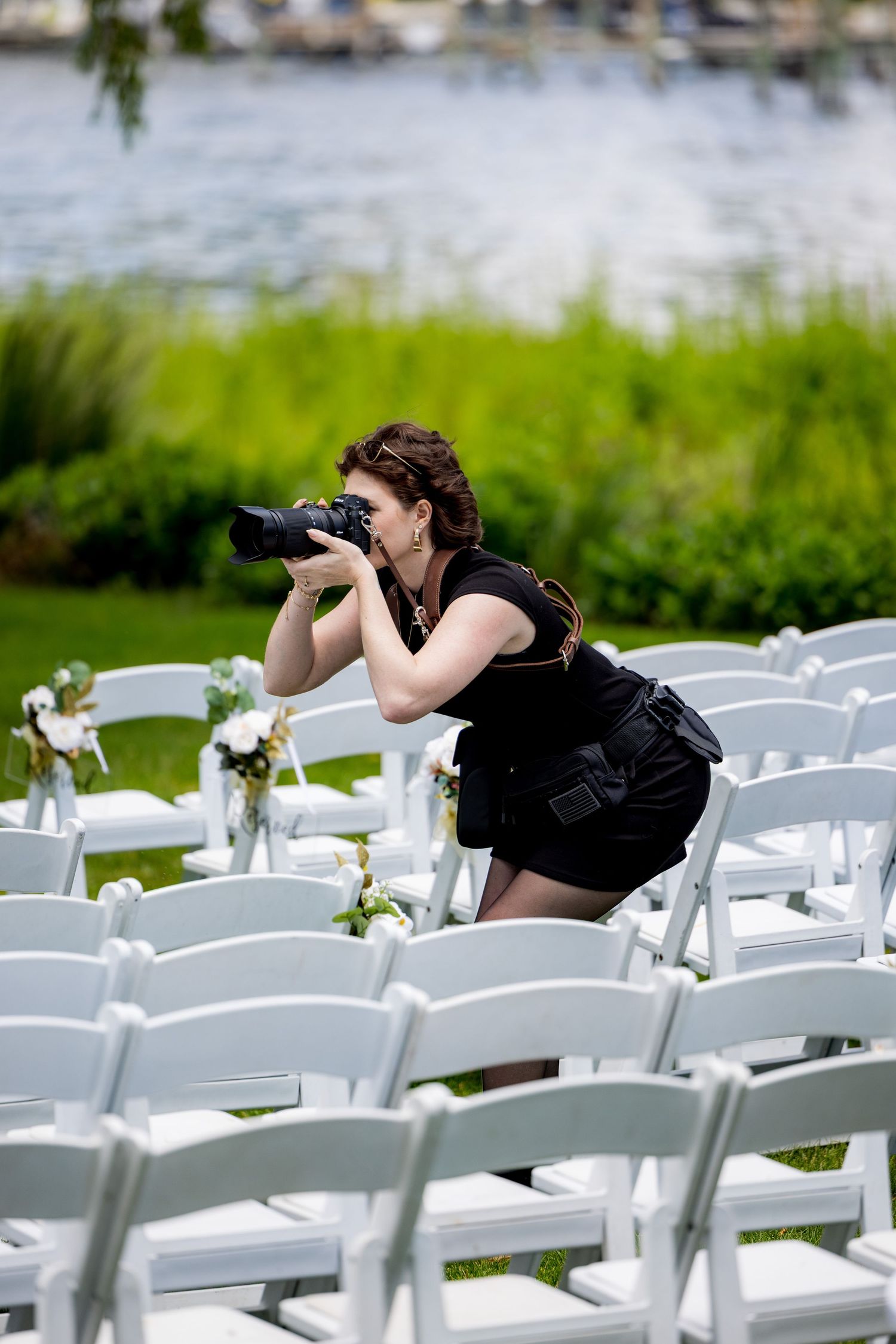 A female wedding photographer crouches among white chairs at an outdoor ceremony, capturing shots with a professional DSLR camera.
