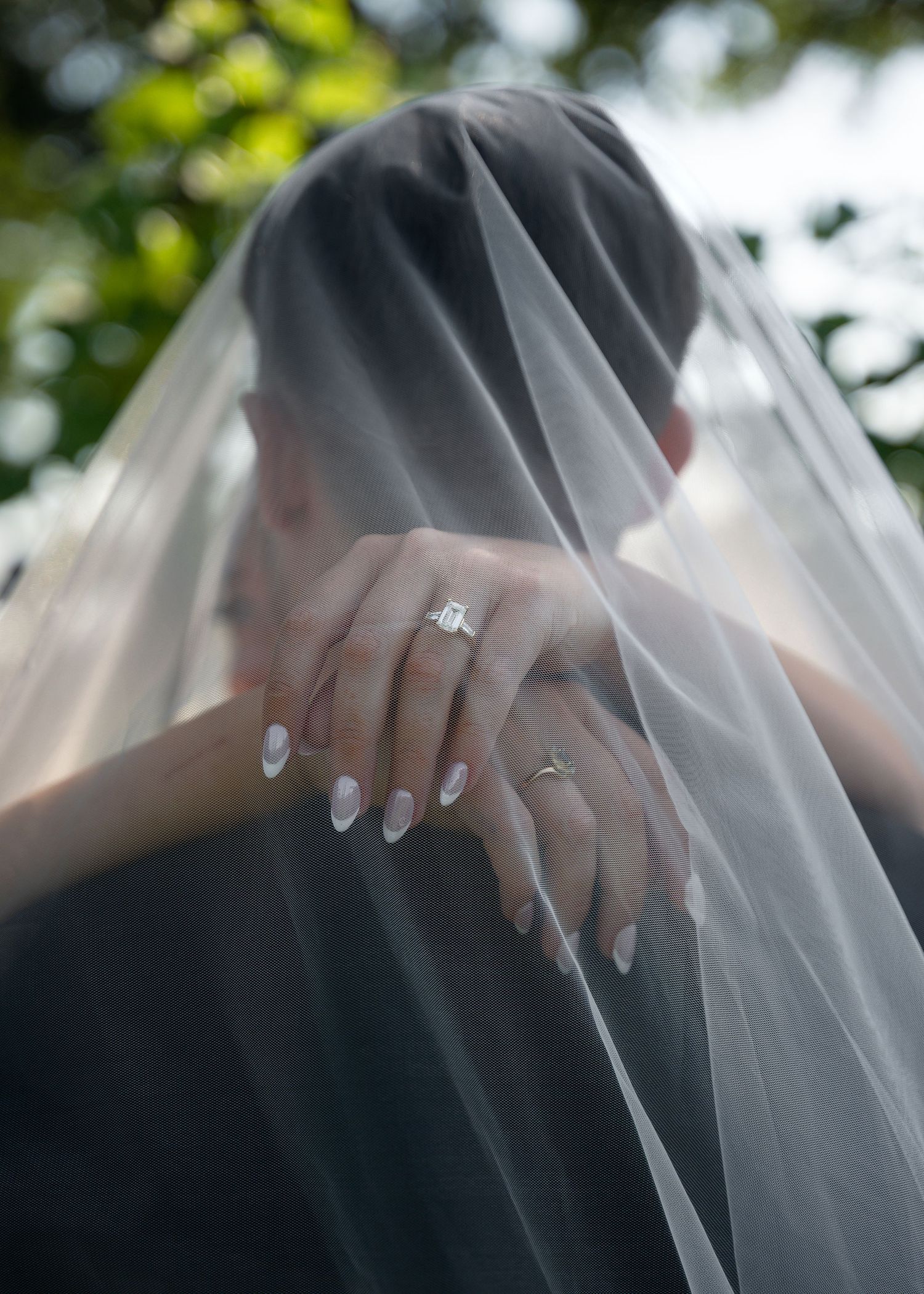 Bride wearing a sheer wedding veil with elegant white nails and a sparkling diamond engagement ring outdoors.