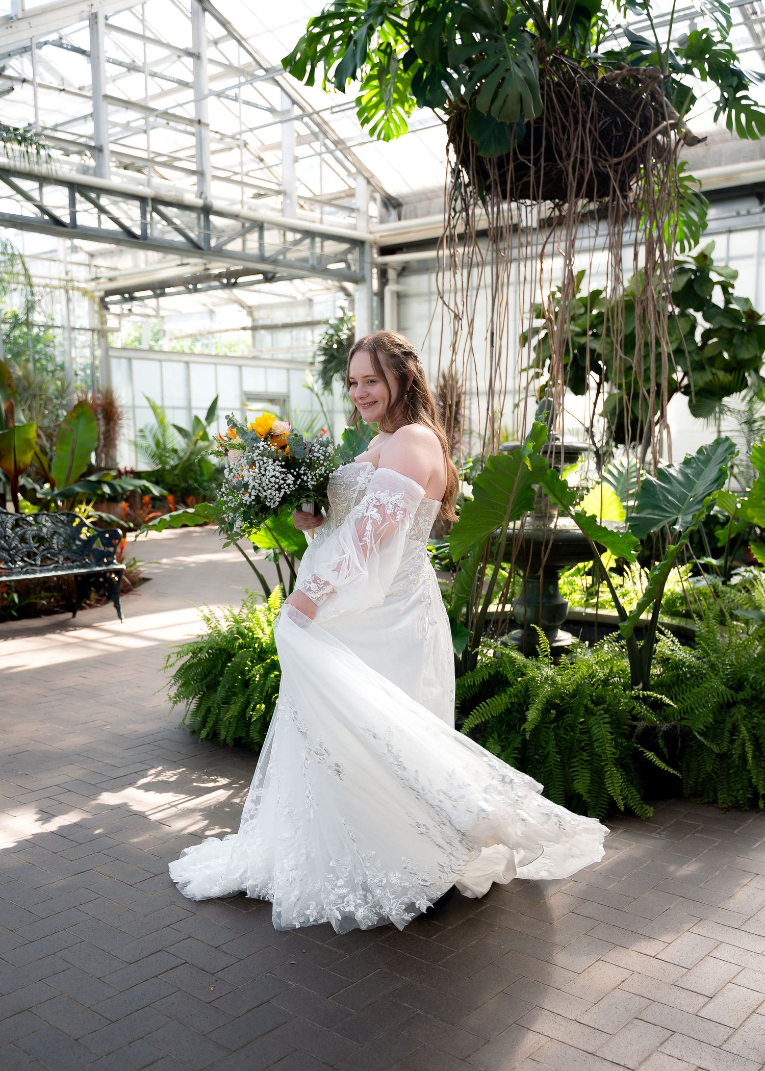 Bride in lace wedding gown holding bouquet smiles in lush greenhouse botanical garden setting.