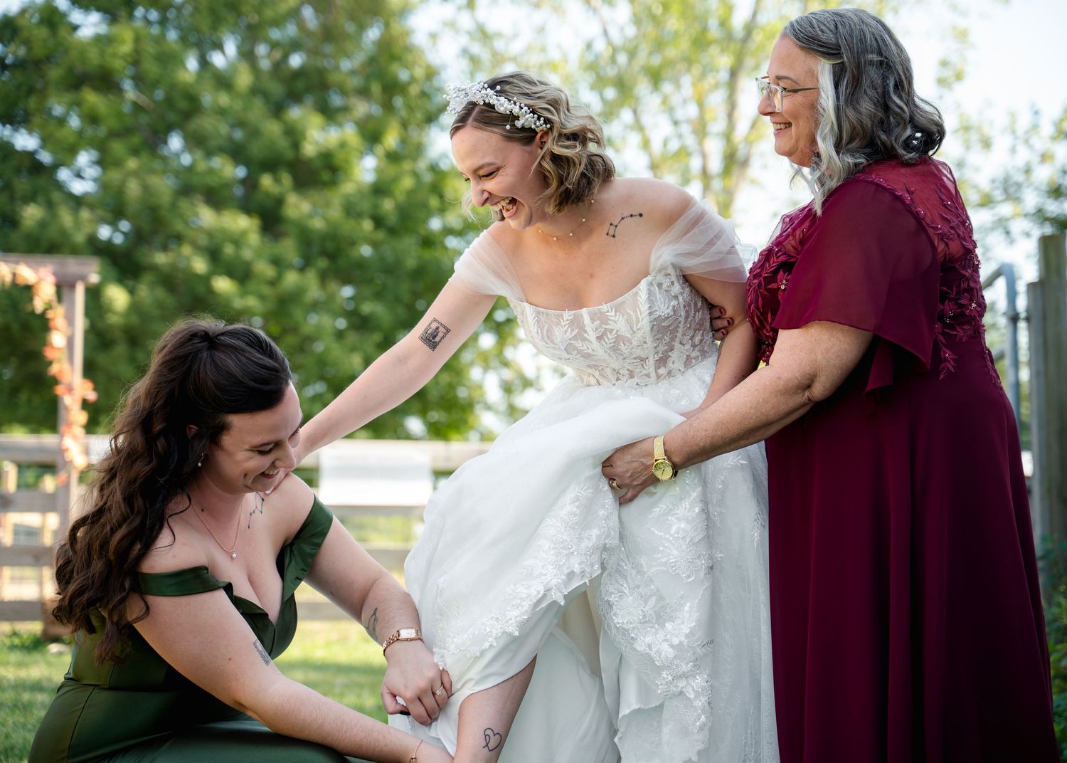 A bride in a lace dress gets help with her gown from attendants in burgundy and black dresses.