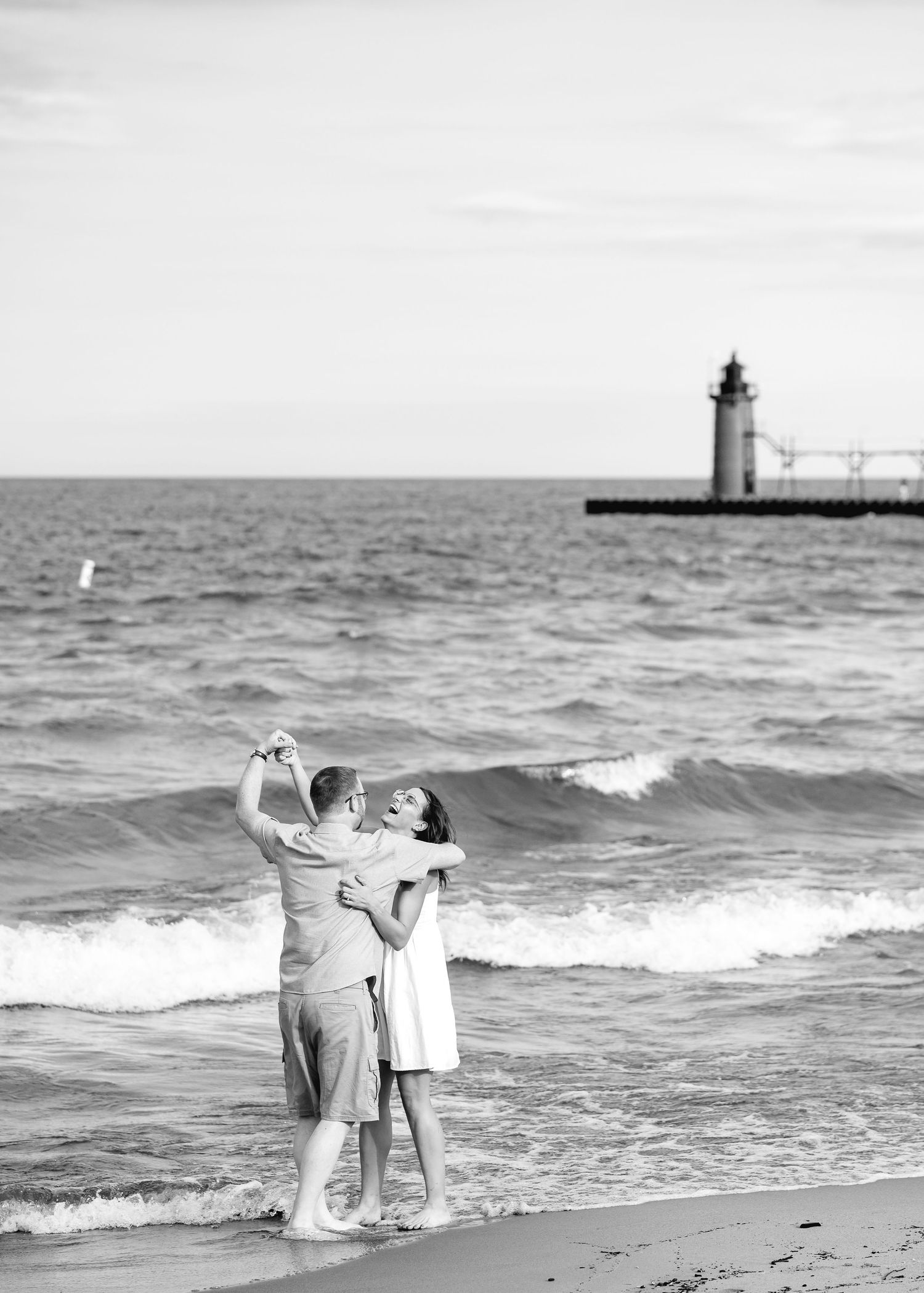 A couple dances romantically on a beach shore in black and white, with a lighthouse pier visible in the background.