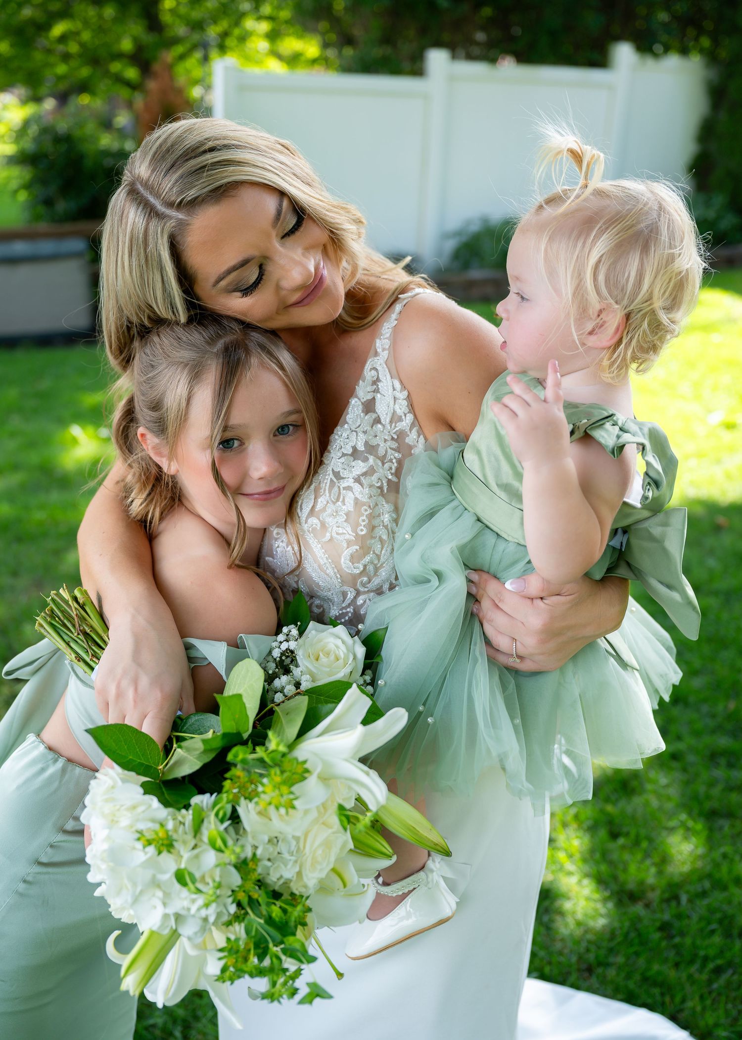 Bride in lace gown hugging two young girls in sage green dresses while holding white floral bouquet outdoors.