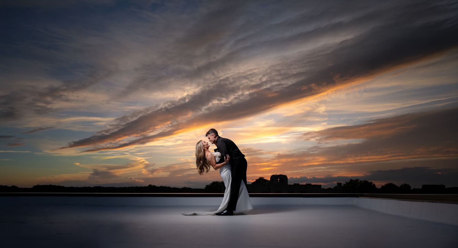 Romantic wedding couple sharing a dip kiss at sunset with dramatic clouds over a serene waterfront backdrop.