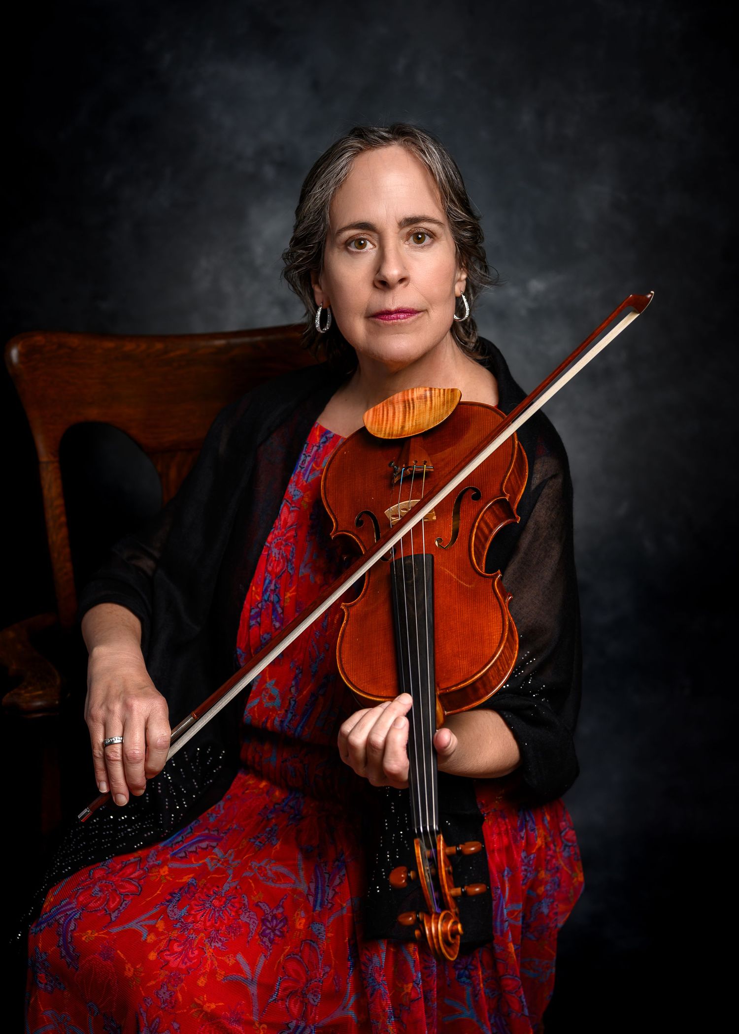 Woman in red floral dress holding violin and bow, seated against dark studio background.