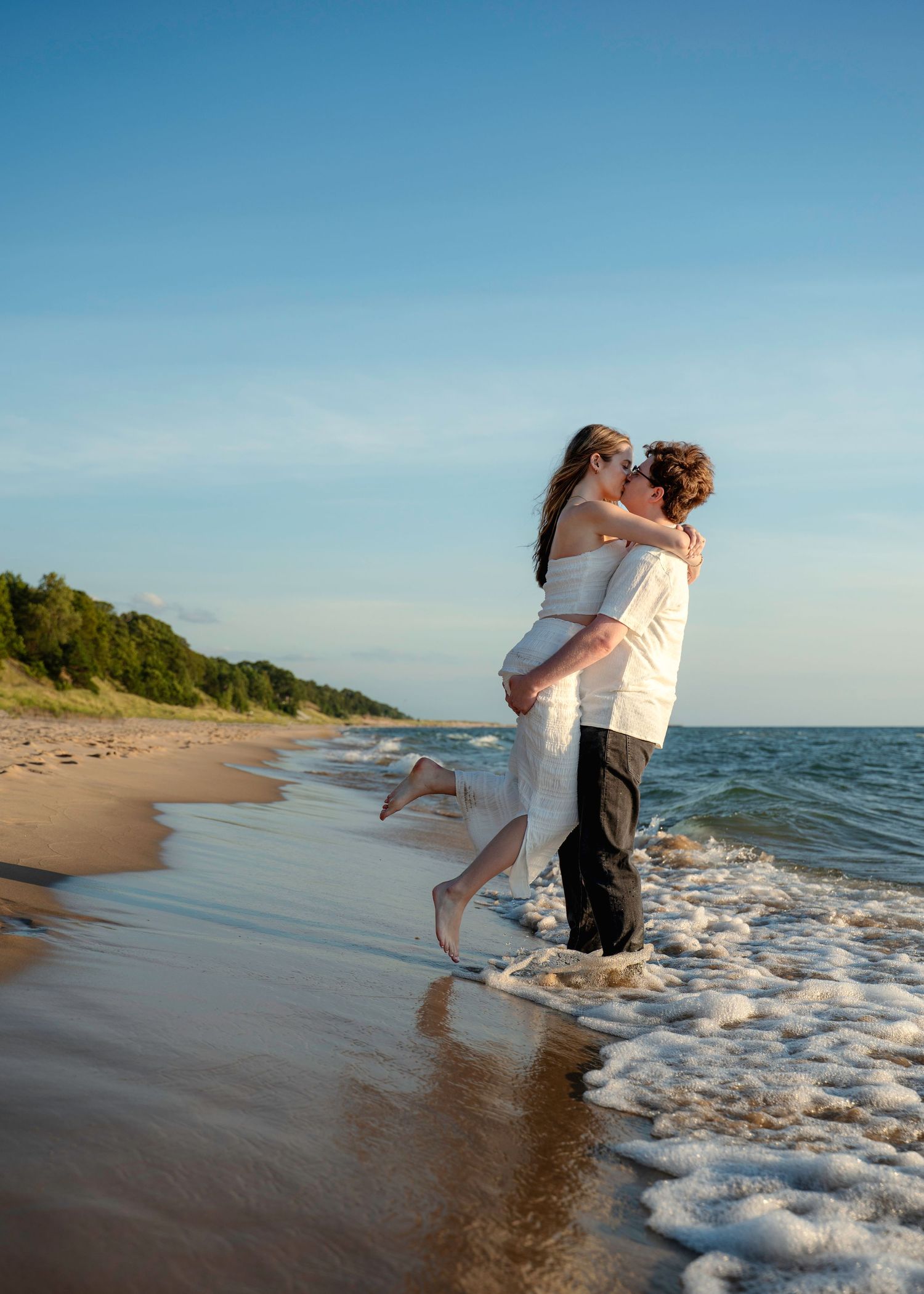Couple in white outfits embracing on beach at sunset with waves crashing.