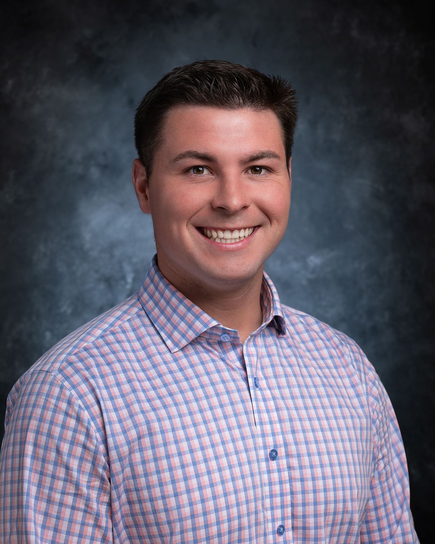 Professional headshot of a smiling young man in a pink plaid shirt against a gray studio background.