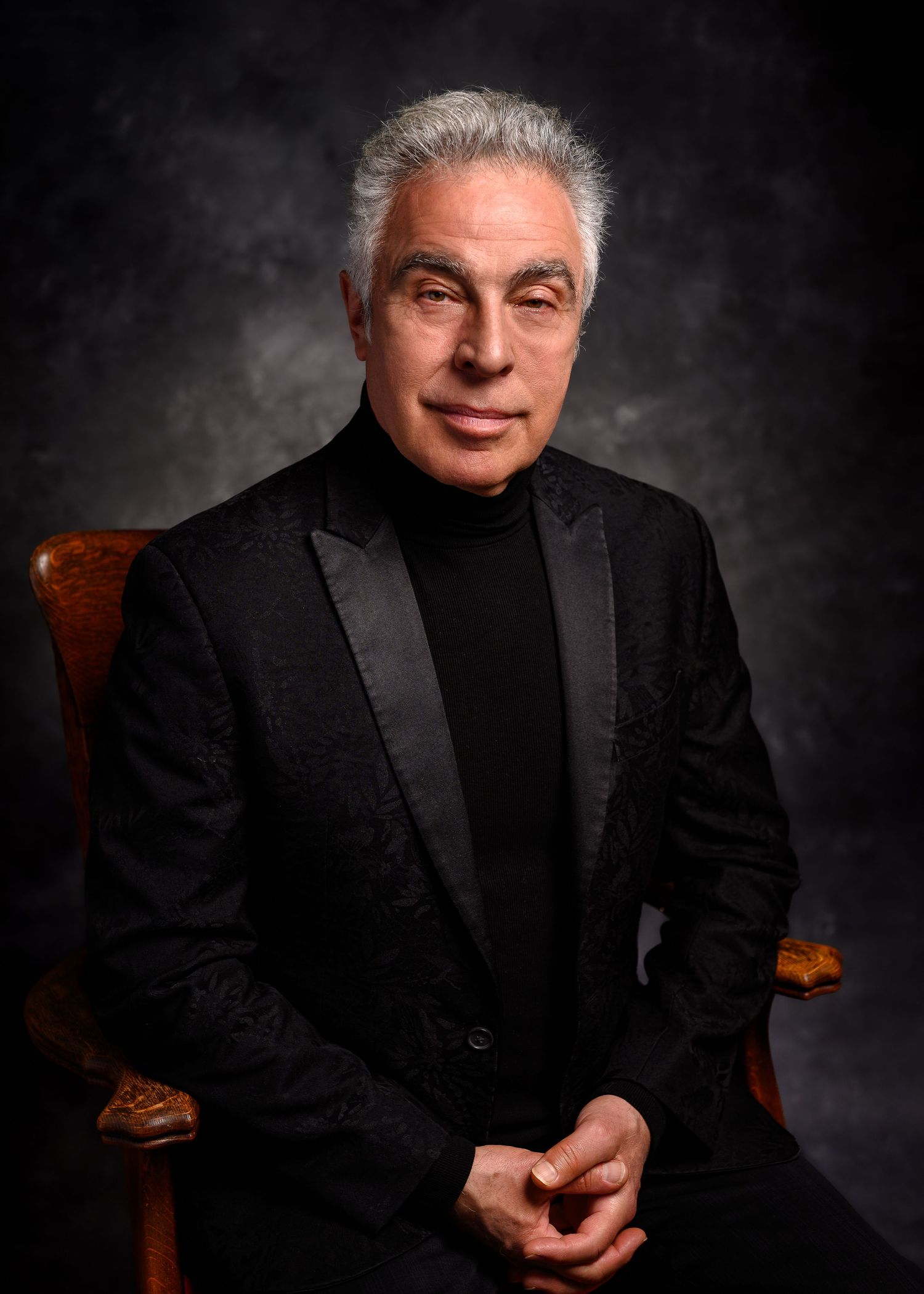 Distinguished older man with silver hair wearing a black turtleneck and blazer, seated in a wooden chair against a dark backdrop.