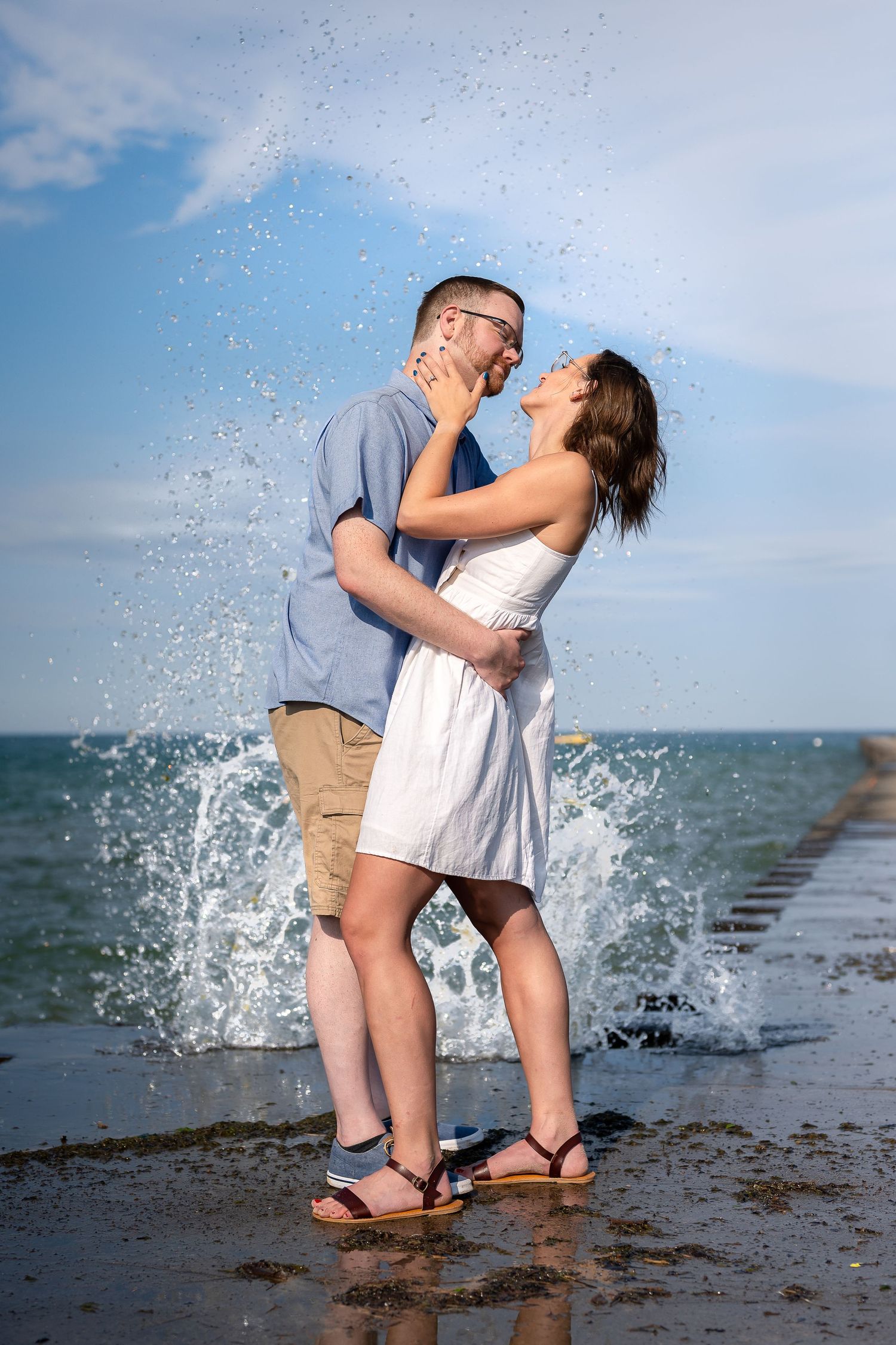 Couple embracing and laughing on a wet beach pier with ocean waves splashing behind them on a sunny day.