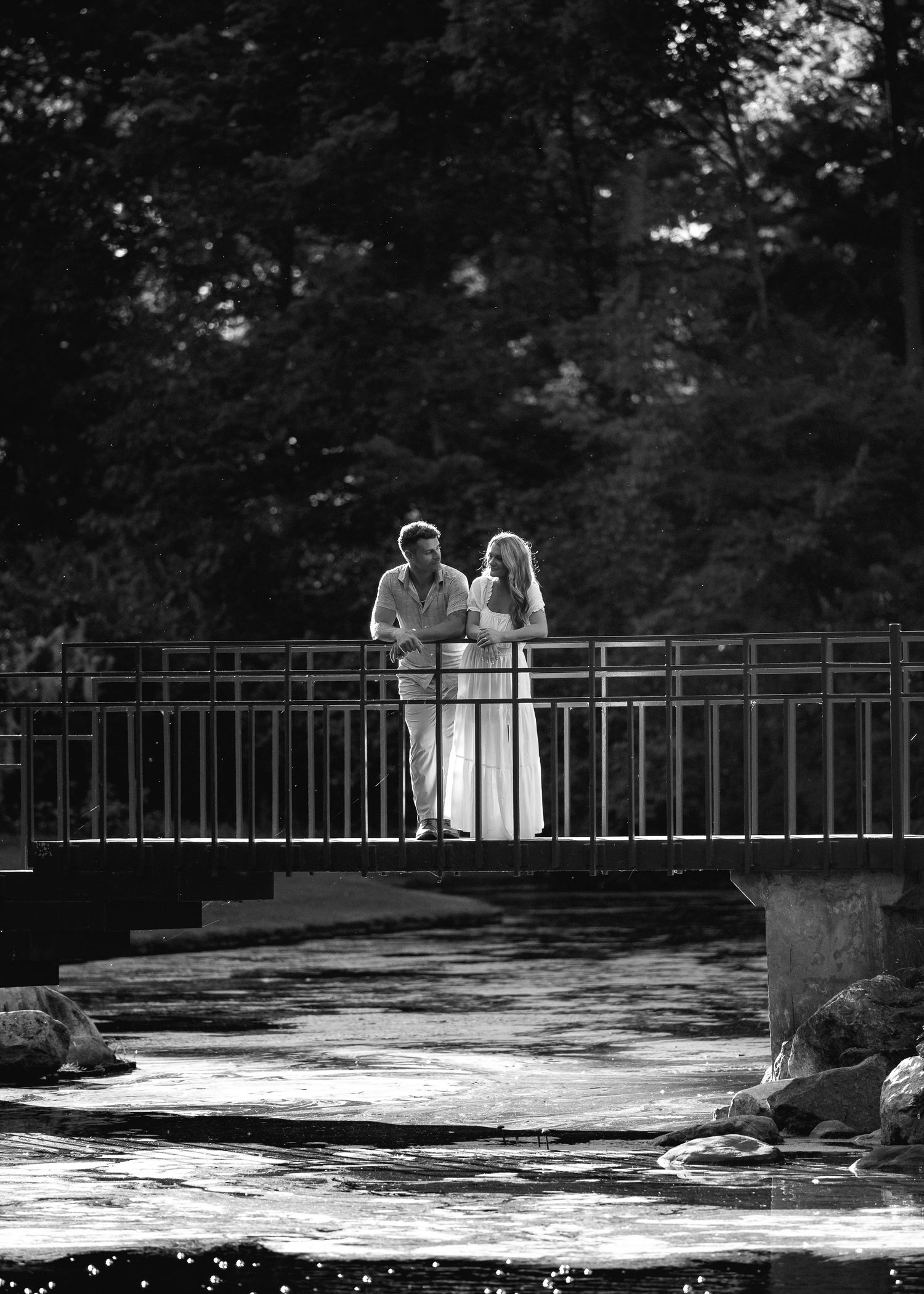 Black and white photo of a couple standing on a bridge surrounded by trees and a rocky stream.