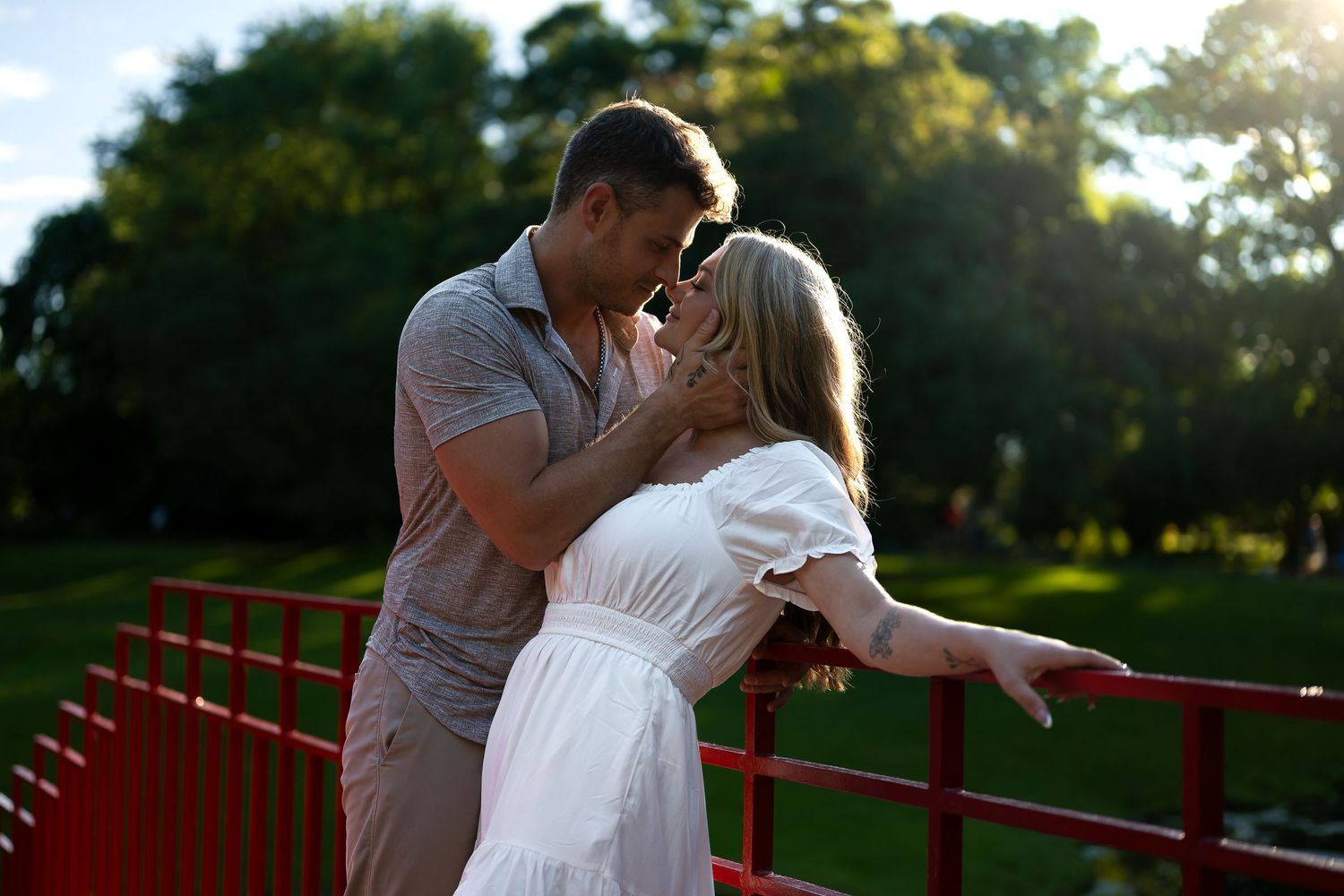 Romantic couple sharing a kiss on a red bridge surrounded by lush green trees in golden sunlight.