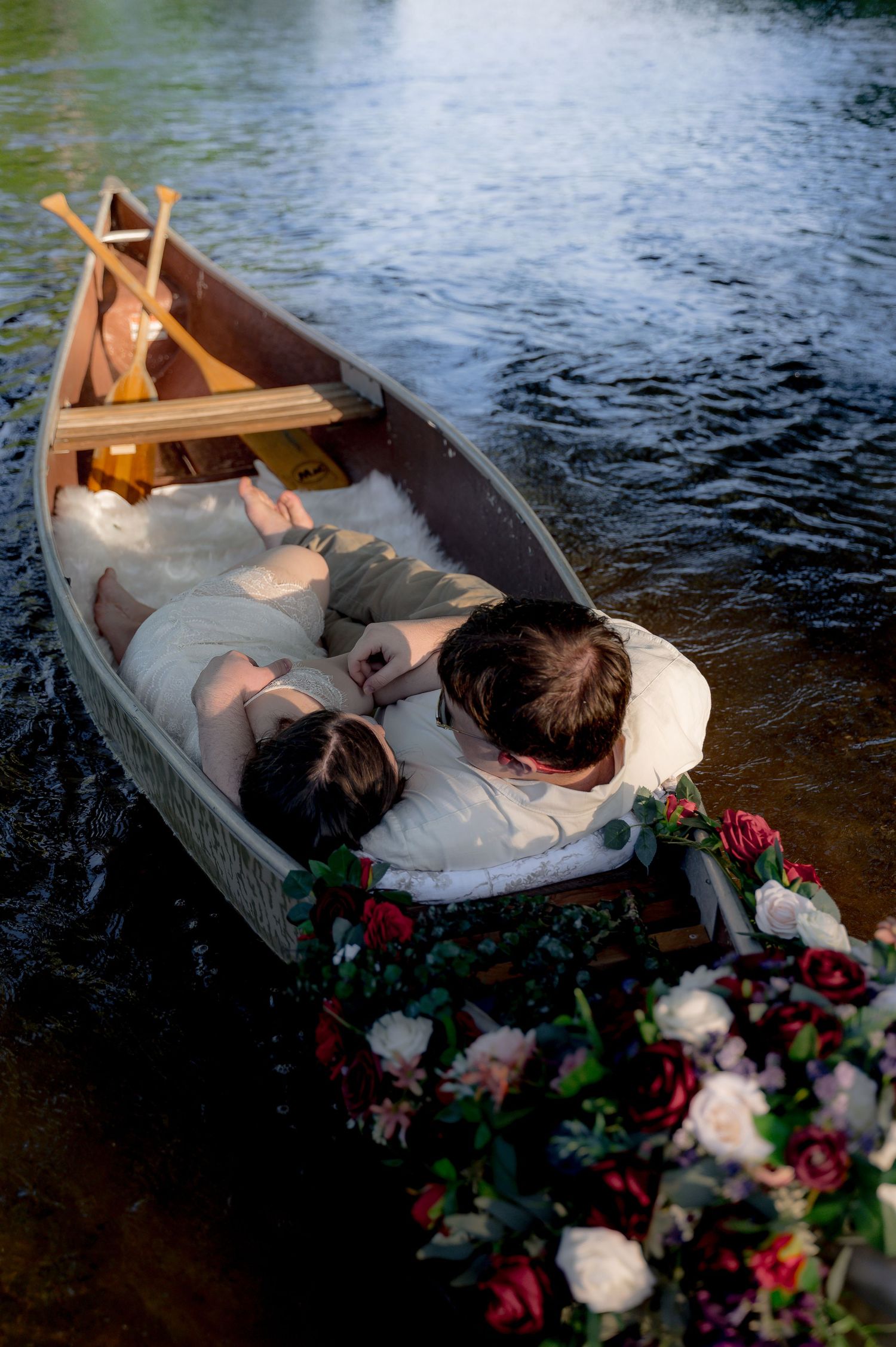 Couple lying in a flower-adorned wooden rowboat on a serene lake, surrounded by lush red and white blooms.