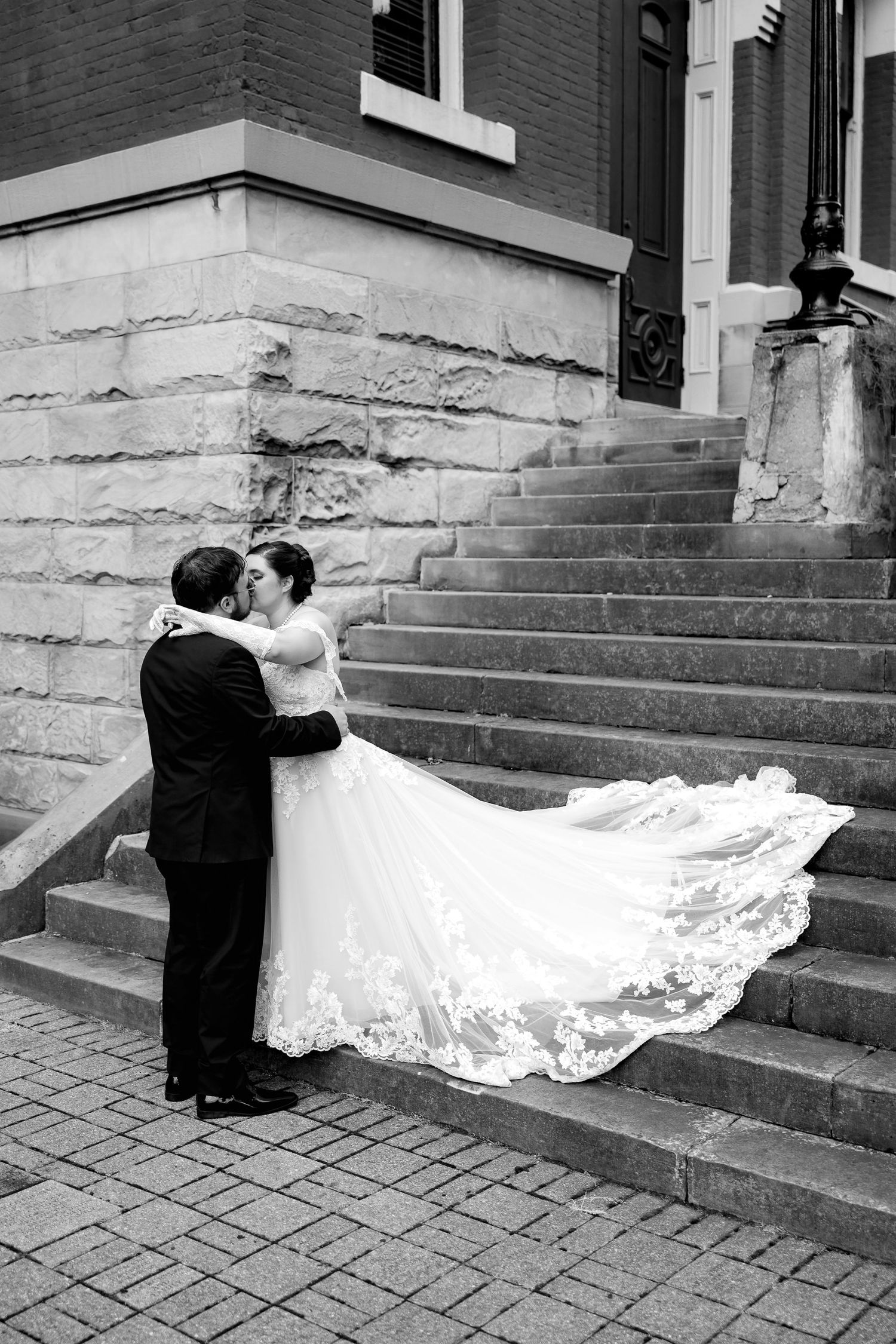 Black and white photo of bride and groom embracing on stone steps, wedding dress train flowing elegantly.