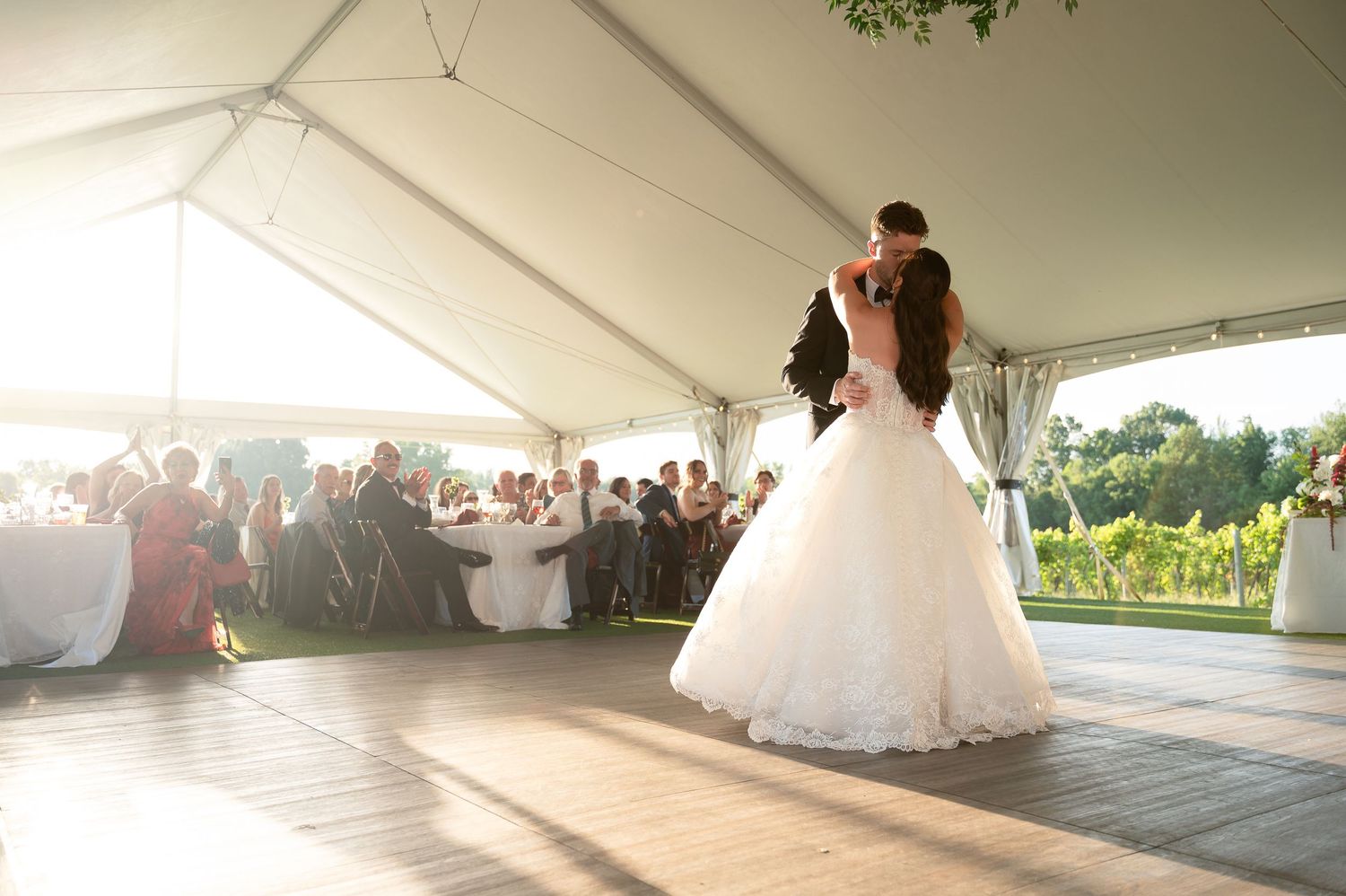 Bride and groom share first dance under elegant white tent at outdoor wedding reception with guests seated nearby.