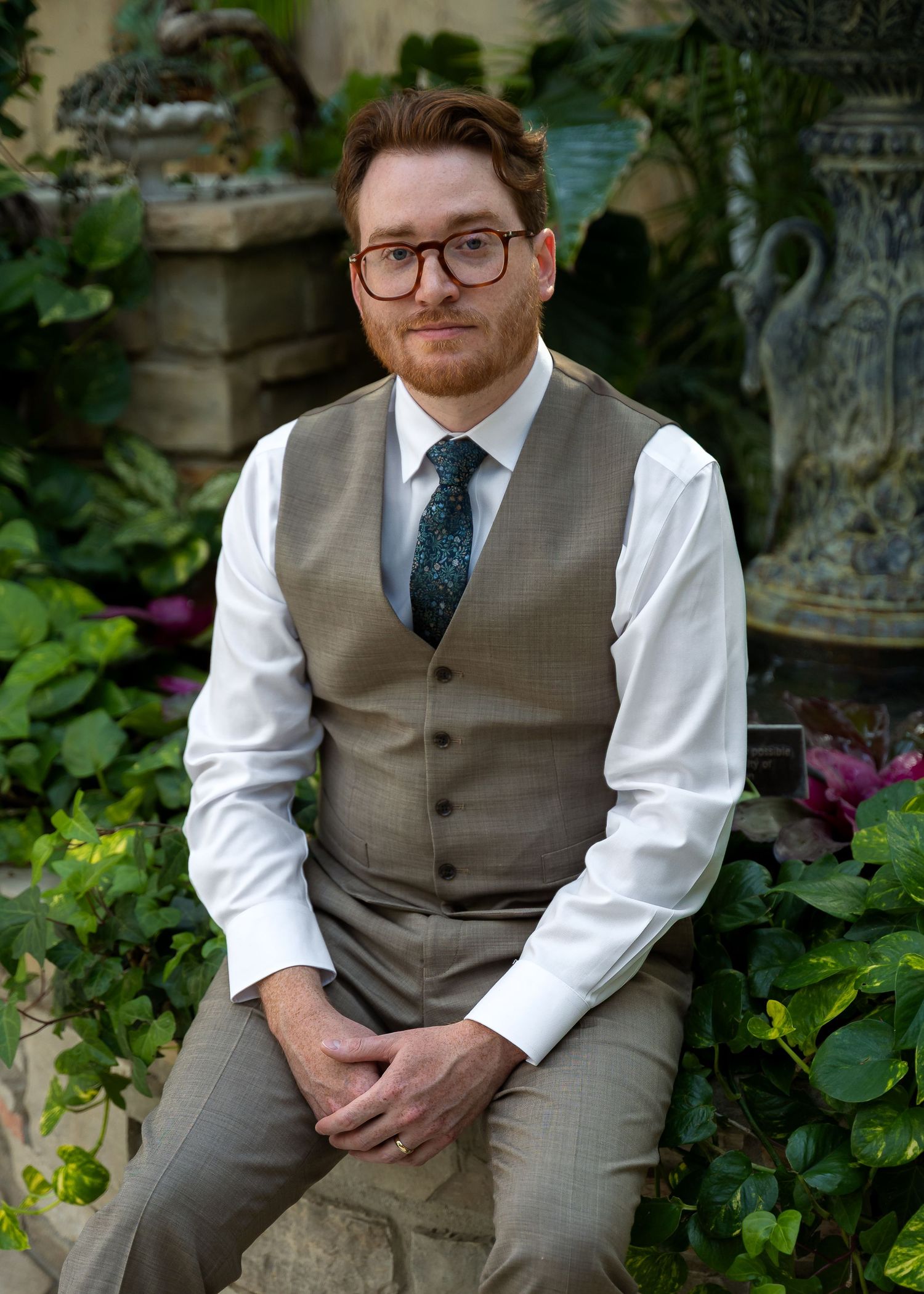 Man in glasses, vest, and tie sits in a lush garden setting, posing for a formal portrait photo.