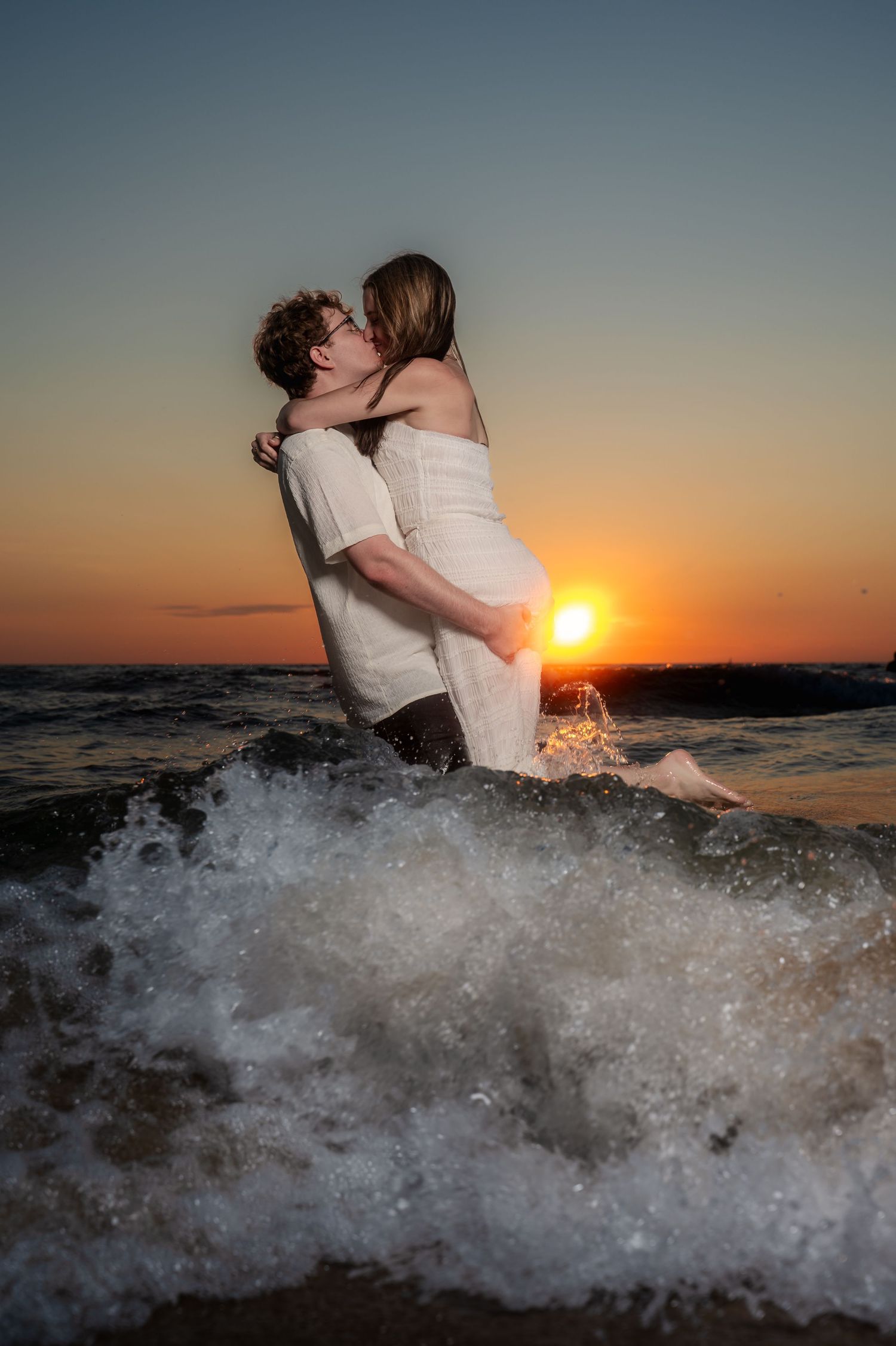 Couple embracing in crashing waves during a dramatic beach sunset.