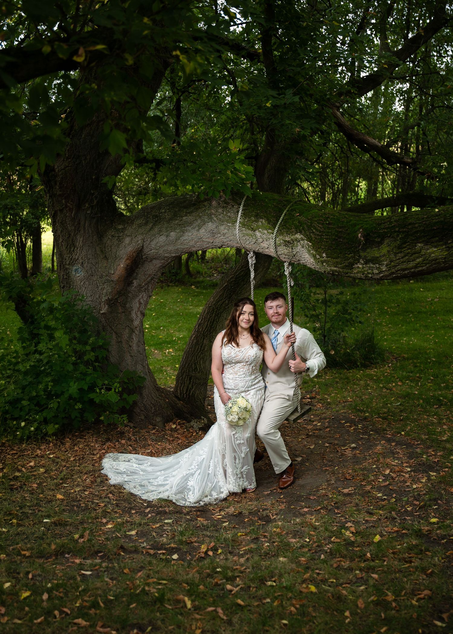 Bride and groom posing by a rustic tree swing in a lush green forest setting on their wedding day.