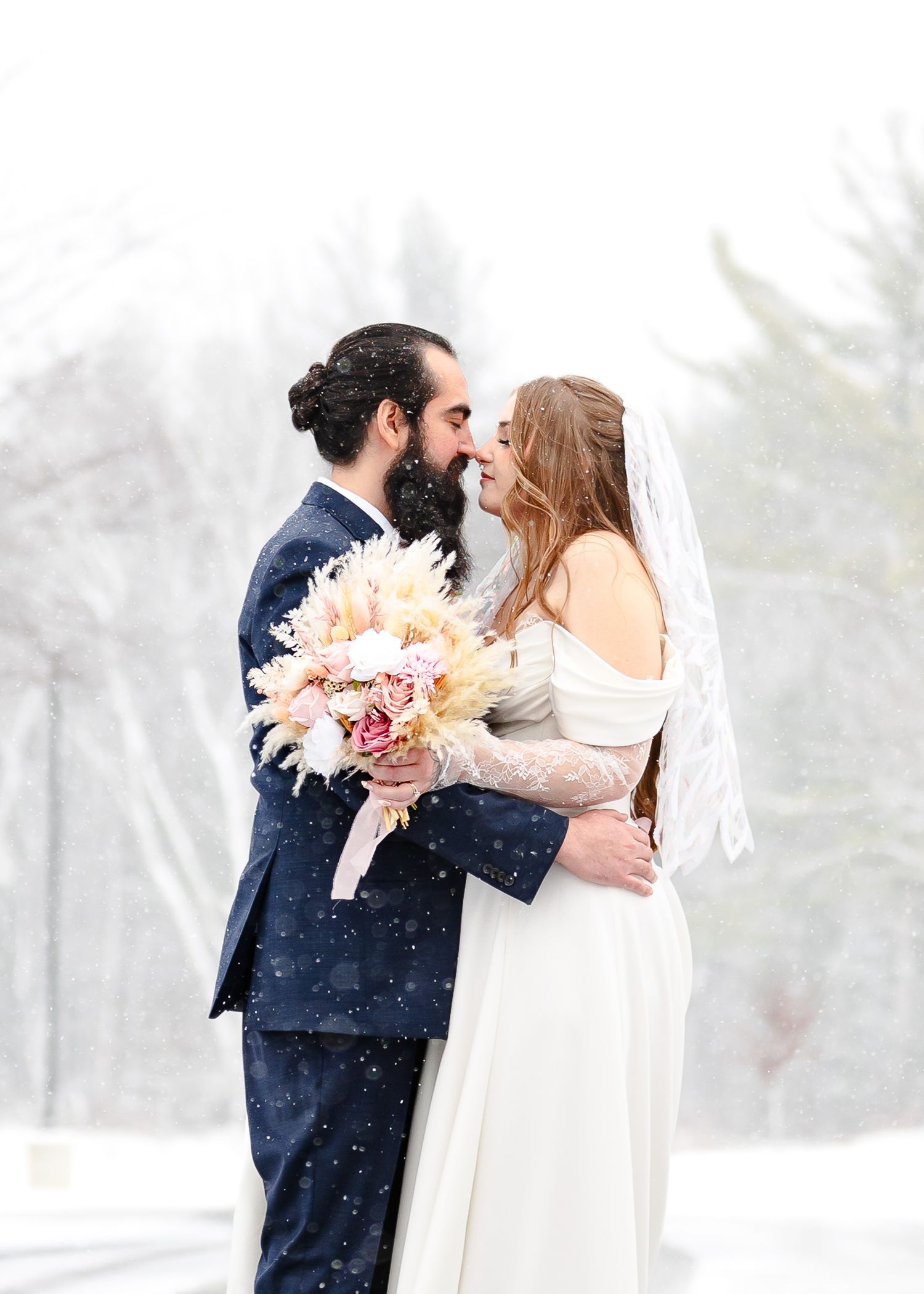 A couple shares a romantic moment in the snow on their winter wedding day, the bride holding a blush bouquet.