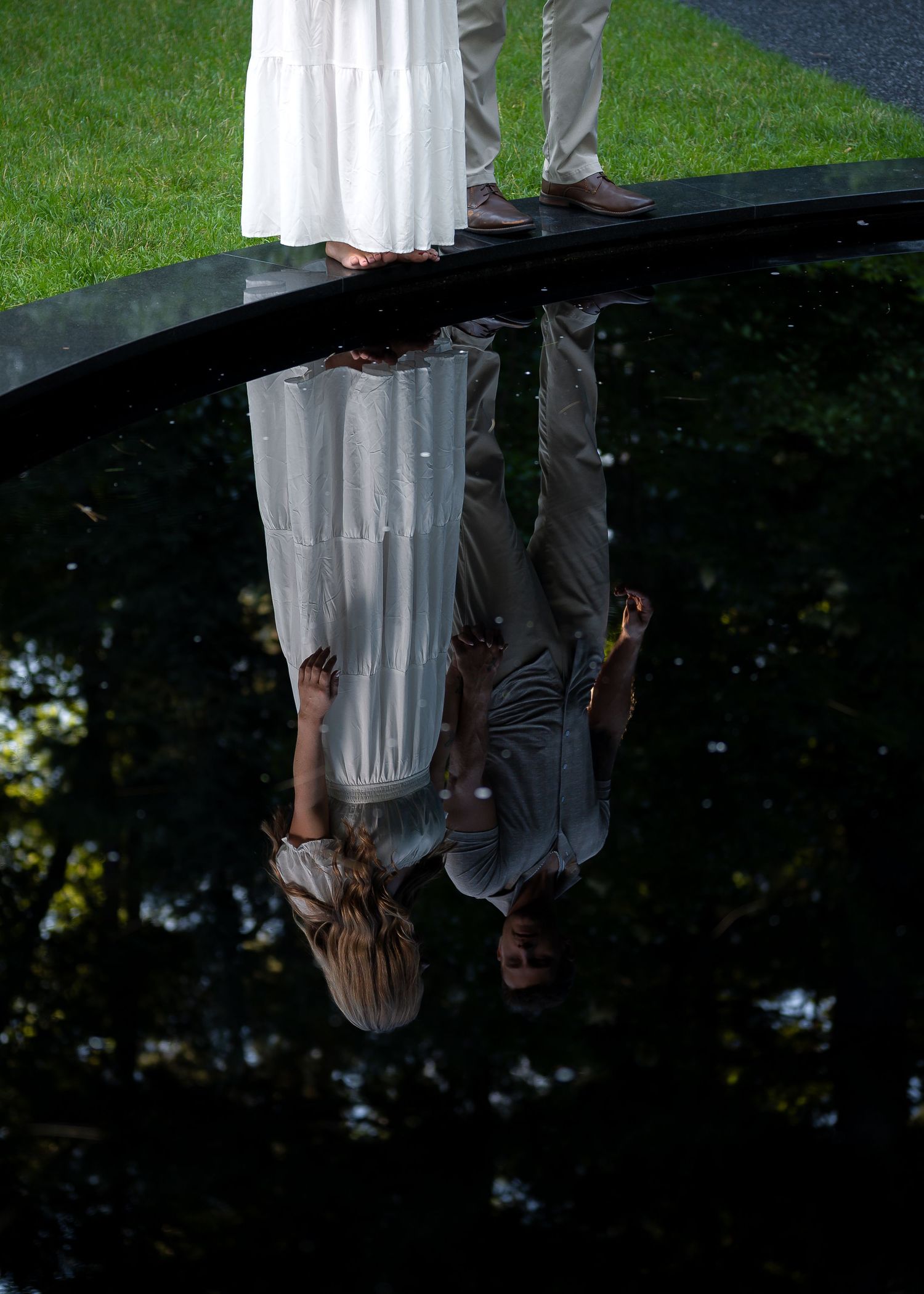 Couple in white dress and gray pants reflected upside down in dark reflective surface, surrounded by lush green trees.