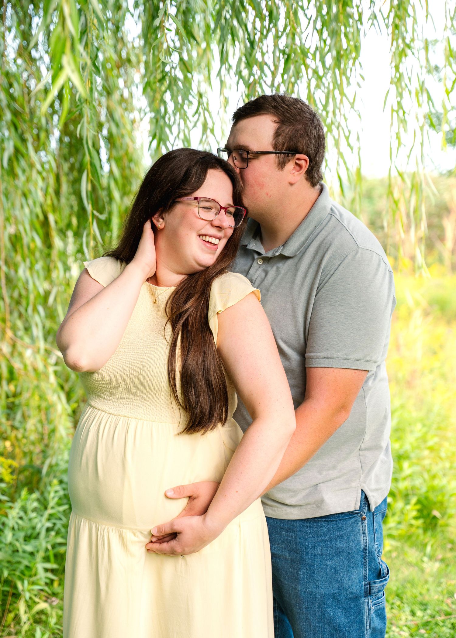 A couple shares an intimate moment under drooping willow branches in natural sunlight.